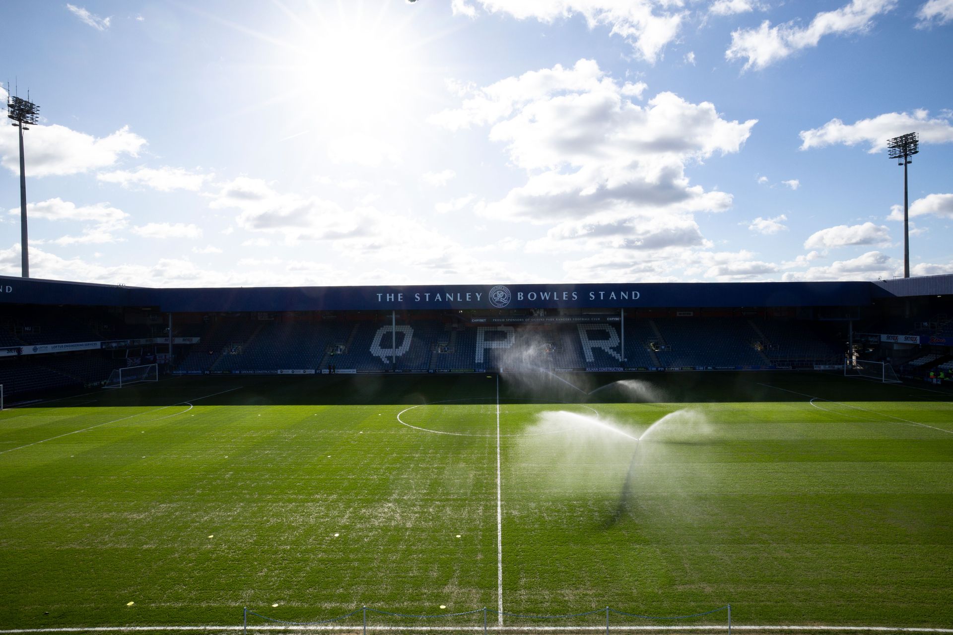 Sunlit soccer stadium with sprinklers watering the green pitch under a blue sky