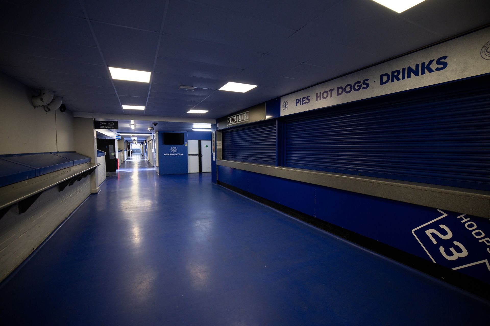 Empty indoor hallway with blue floor, closed concession shutters, and signs for hot dogs and drinks