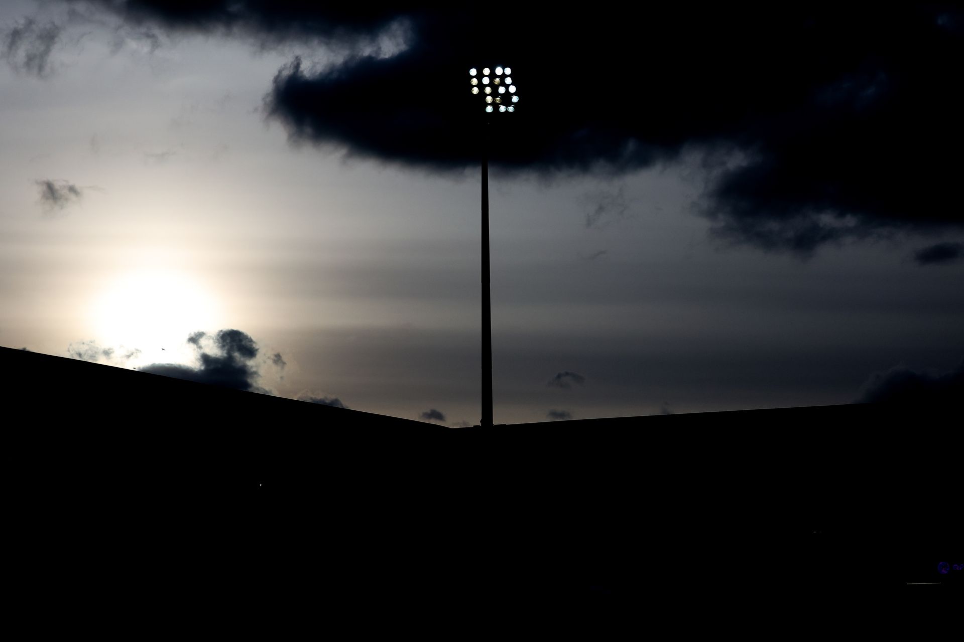 Silhouetted stadium light pole at dusk with dark clouds and a bright sky on the horizon