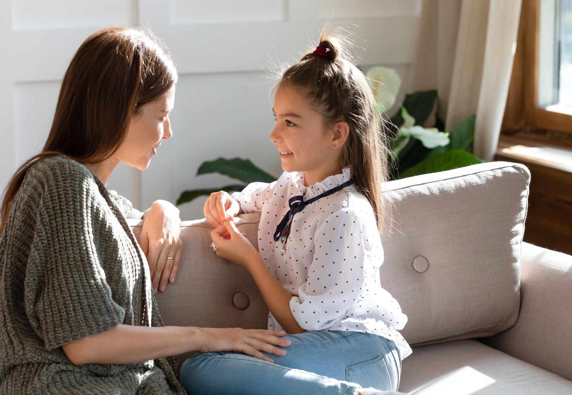 Woman and girl seated on a couch, talking. Girl smiles, holding an object. Bright, indoor setting.