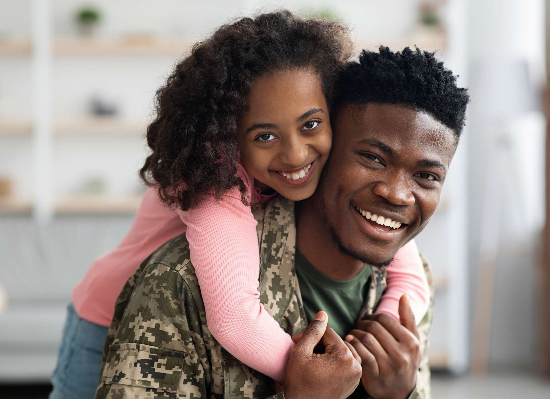Smiling person in camouflage uniform is embraced by a smiling girl; indoor setting. Smiling person in camouflage uniform is embraced by a smiling girl; indoor setting.