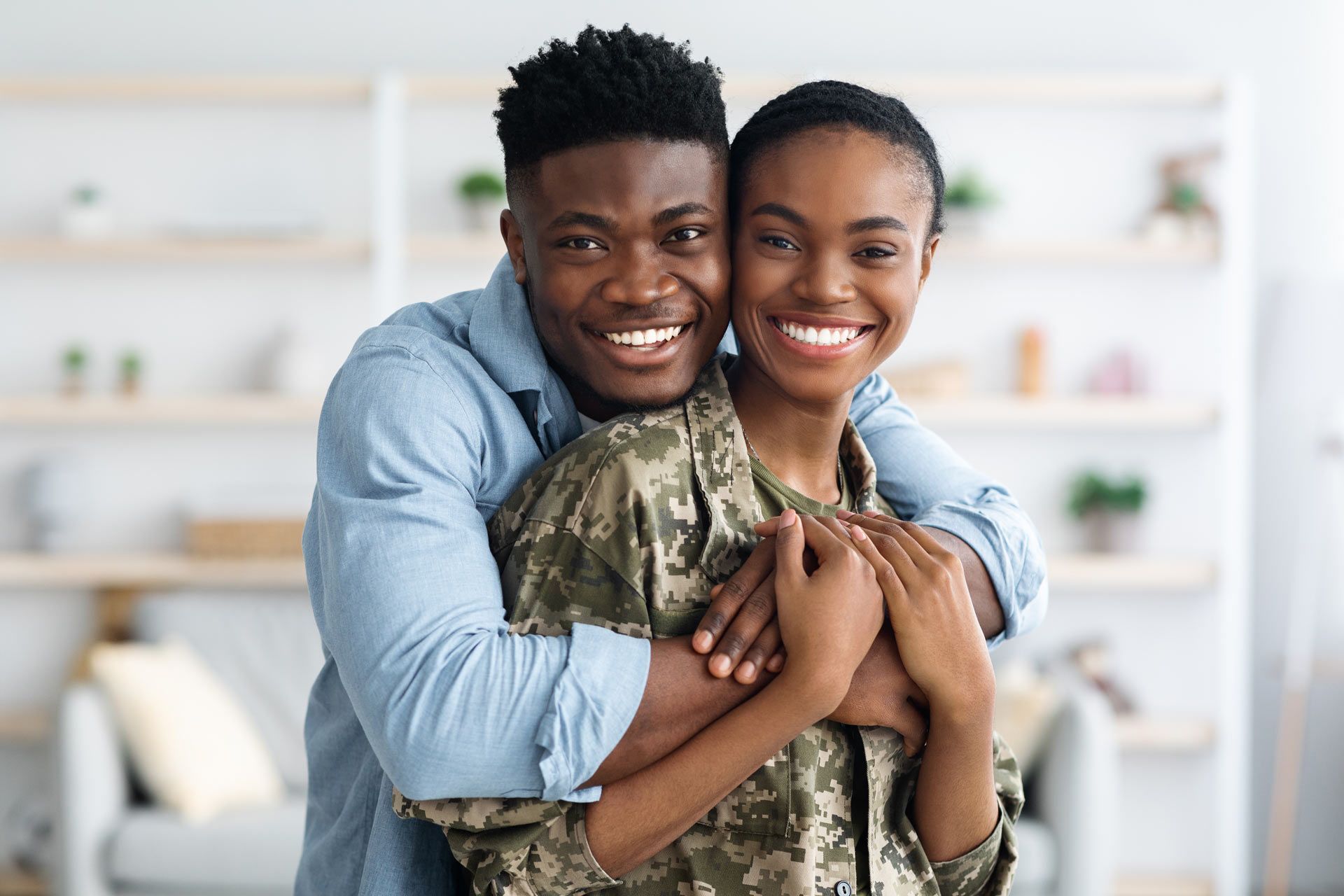 Man hugs woman in camouflage; both smile indoors.