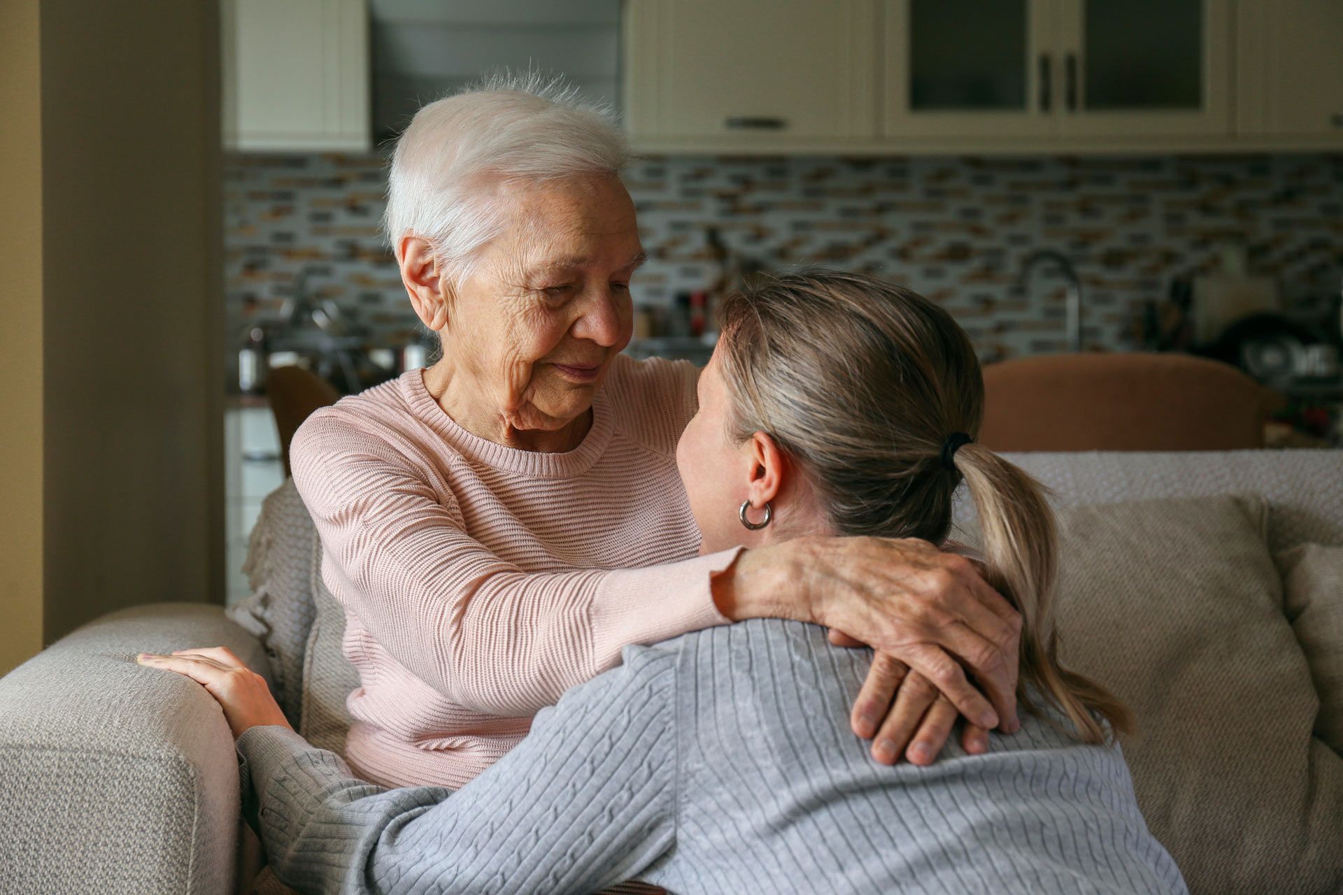 Older woman embraces another woman on a couch; indoor setting.