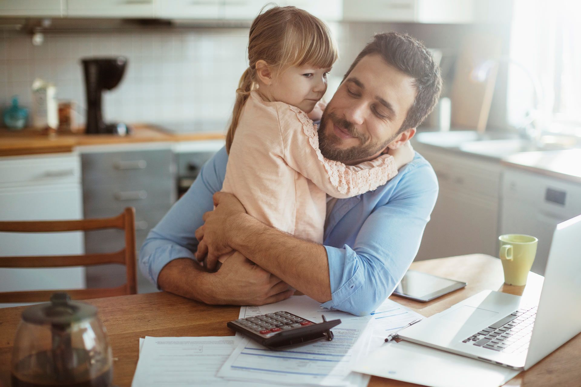 Man hugging a child while working at a table with papers, calculator, and laptop in a kitchen.
