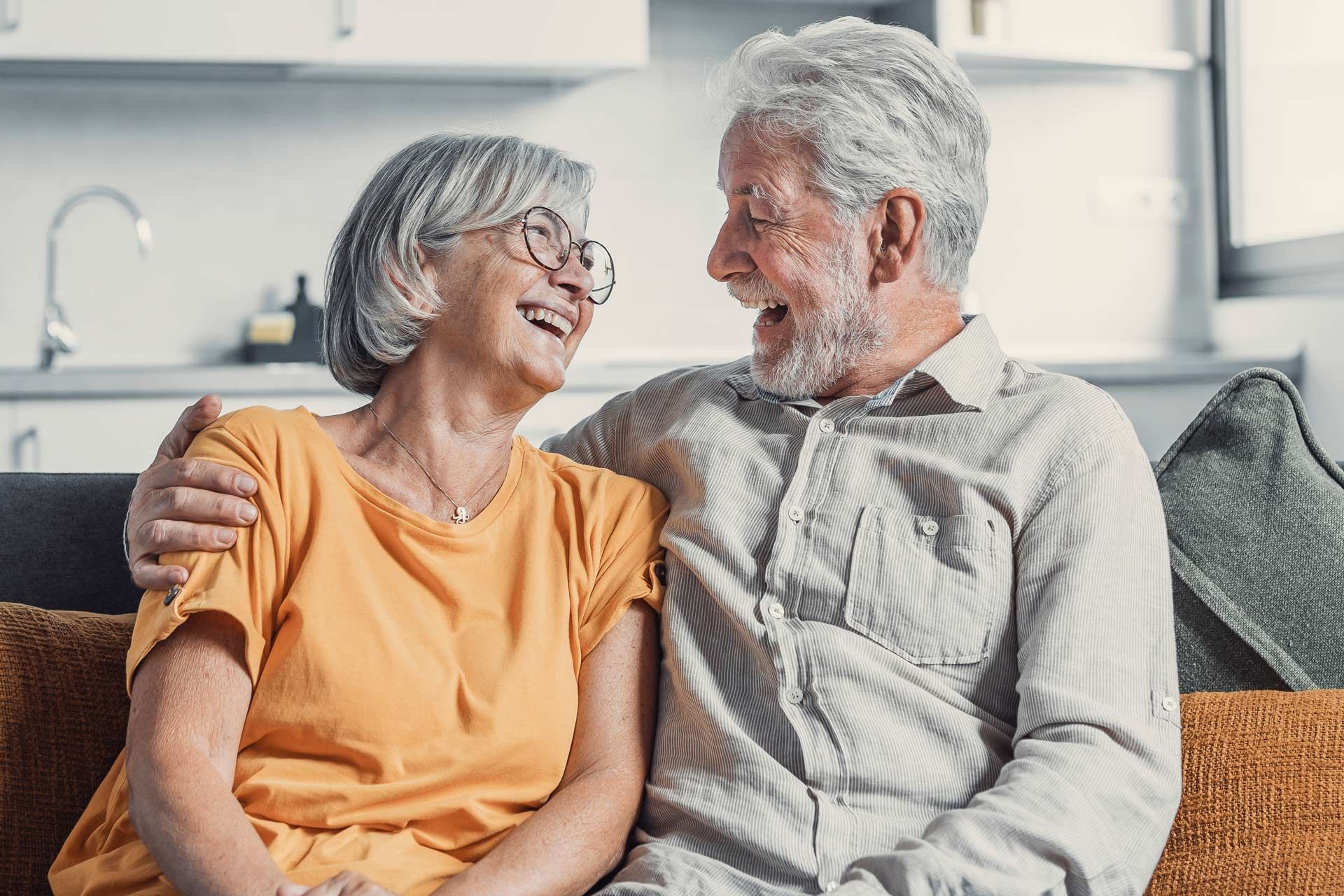 An older couple smiles, seated on a couch indoors. Man has arm around woman.