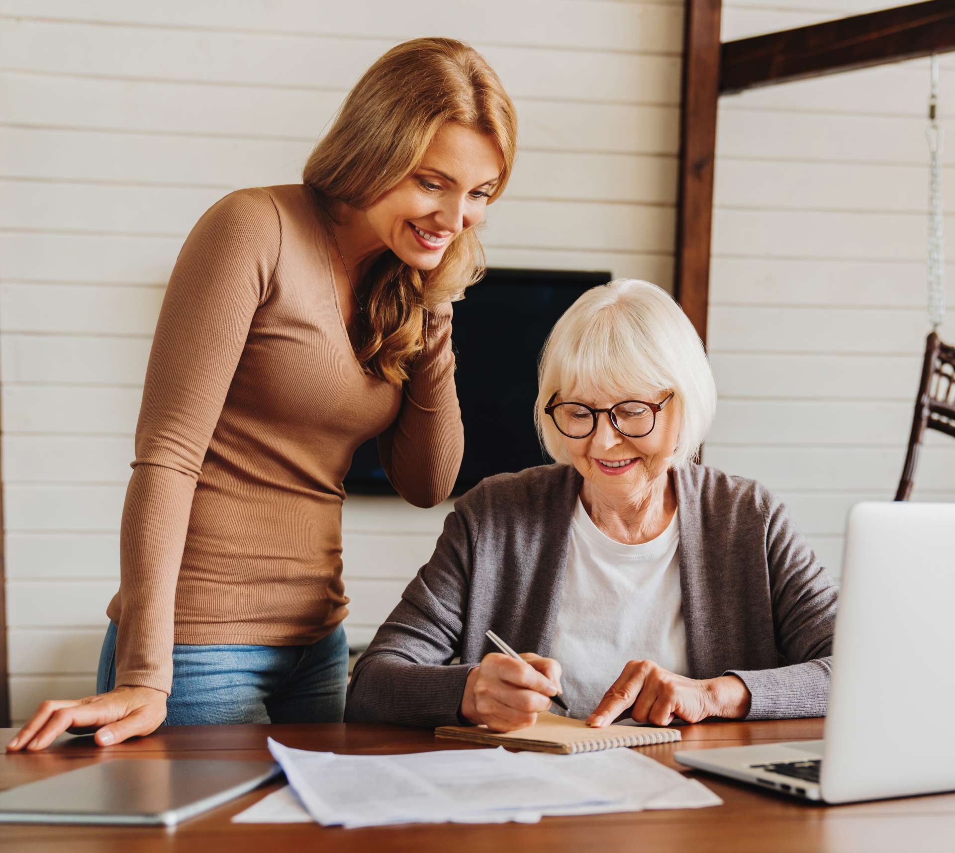 Woman assisting an older person with paperwork, both smiling, laptop and papers on a table.