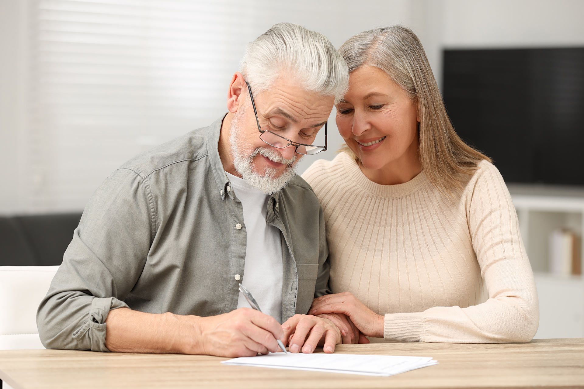 Senior couple at table, man writing on paper, woman looking on with smile. Indoors, neutral colors.