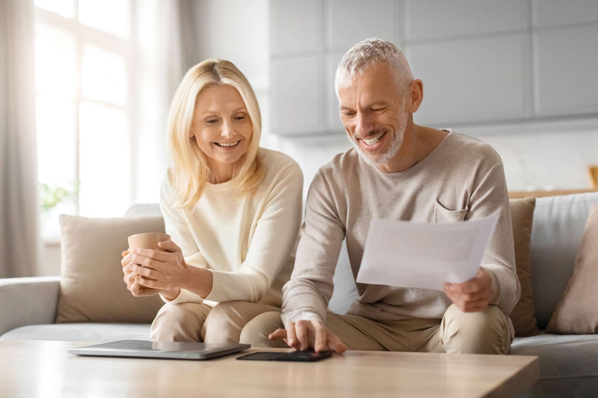 Couple reviewing documents, smiling, sitting on a sofa with a tablet and calculator.