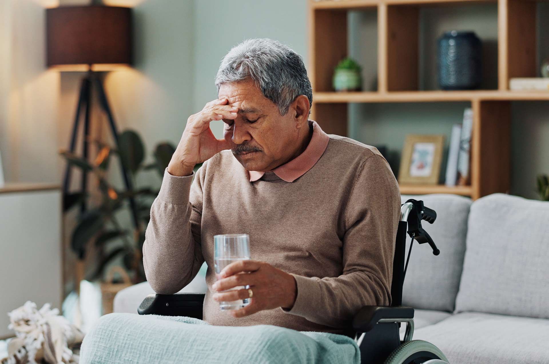 Older person in a wheelchair, holding a glass of water, and touching his forehead indoors, looking distressed.