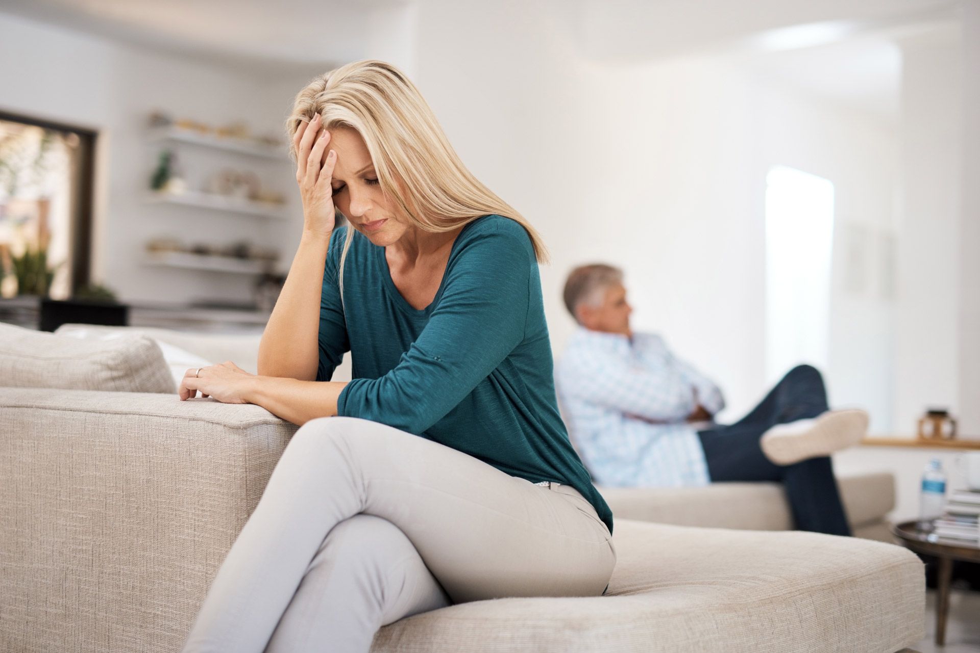Woman with head in hand sits on couch; man sits behind her, both appear distressed in a living room.