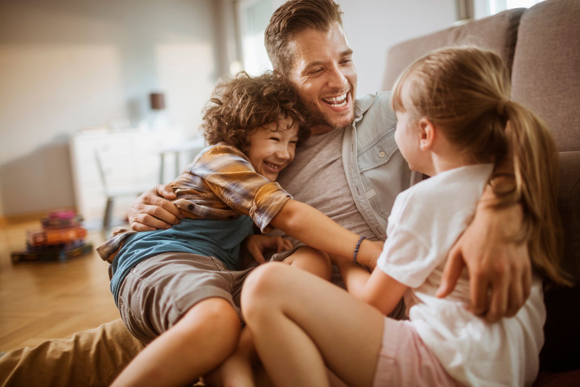 Man smiles, embracing two children on a couch. They laugh, appearing happy in a living room setting.