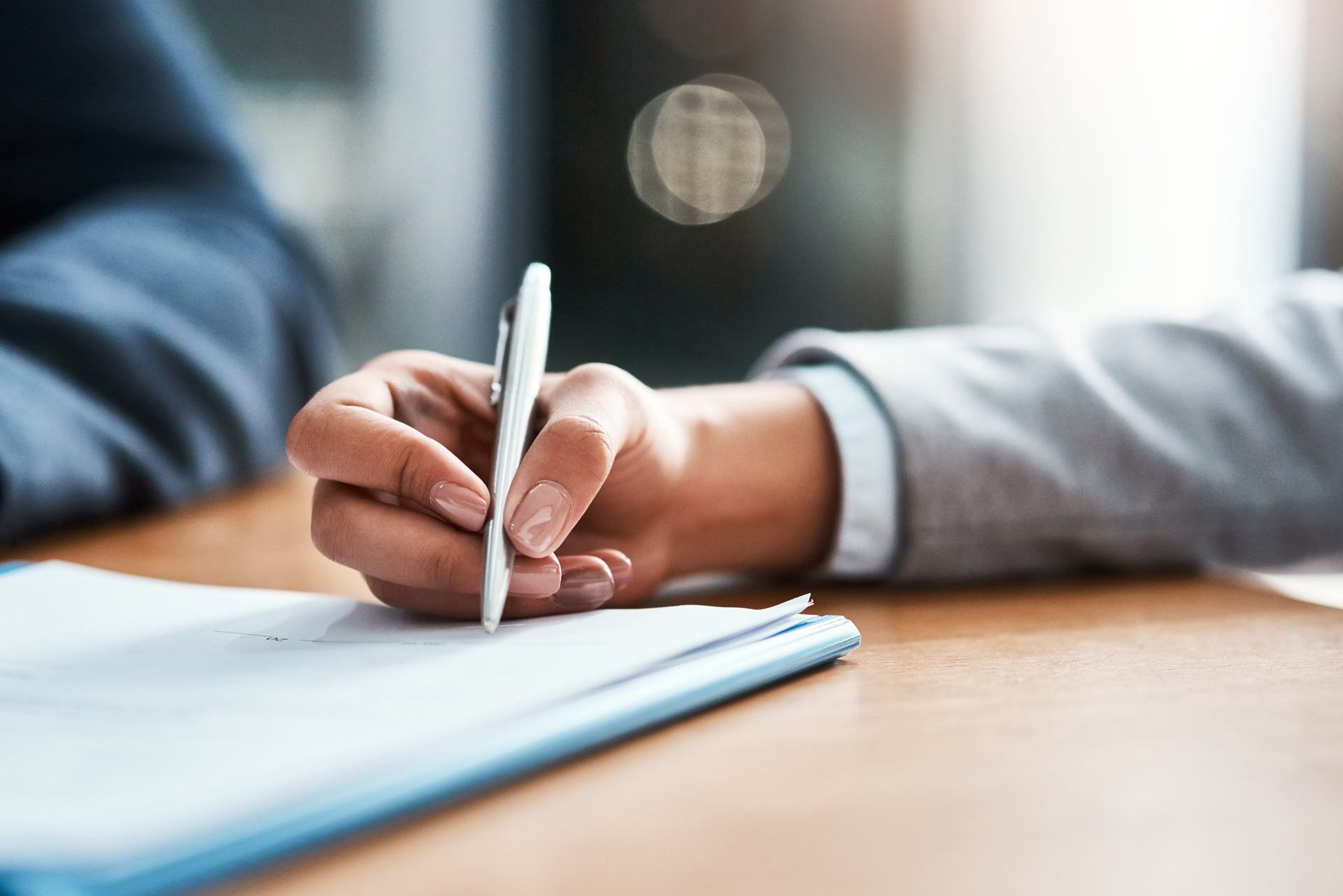 Person in a grey blazer writing on a document with a silver pen. The document rests on a clipboard.