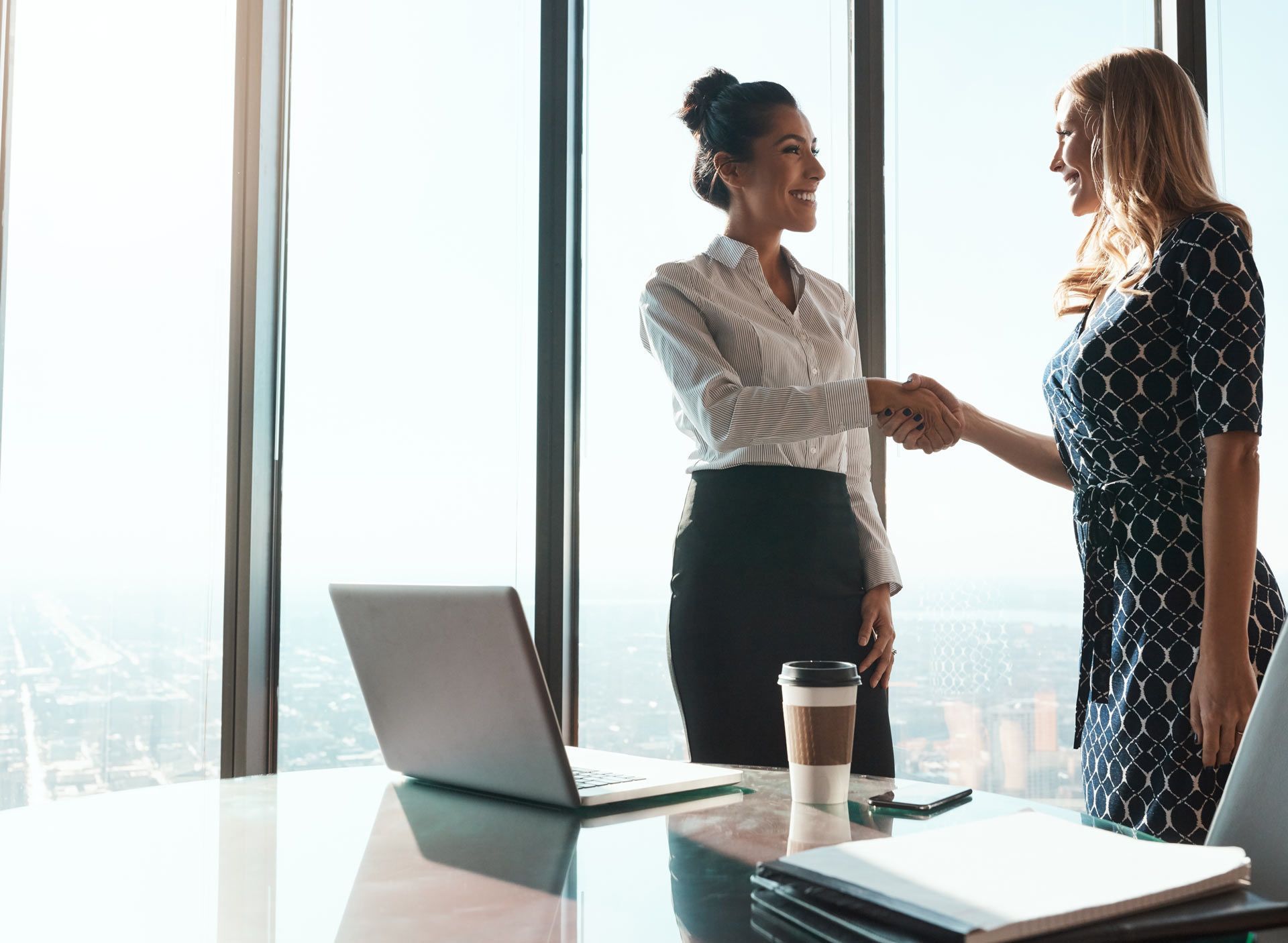 Two women in business attire shaking hands in a bright office, cityscape visible outside.