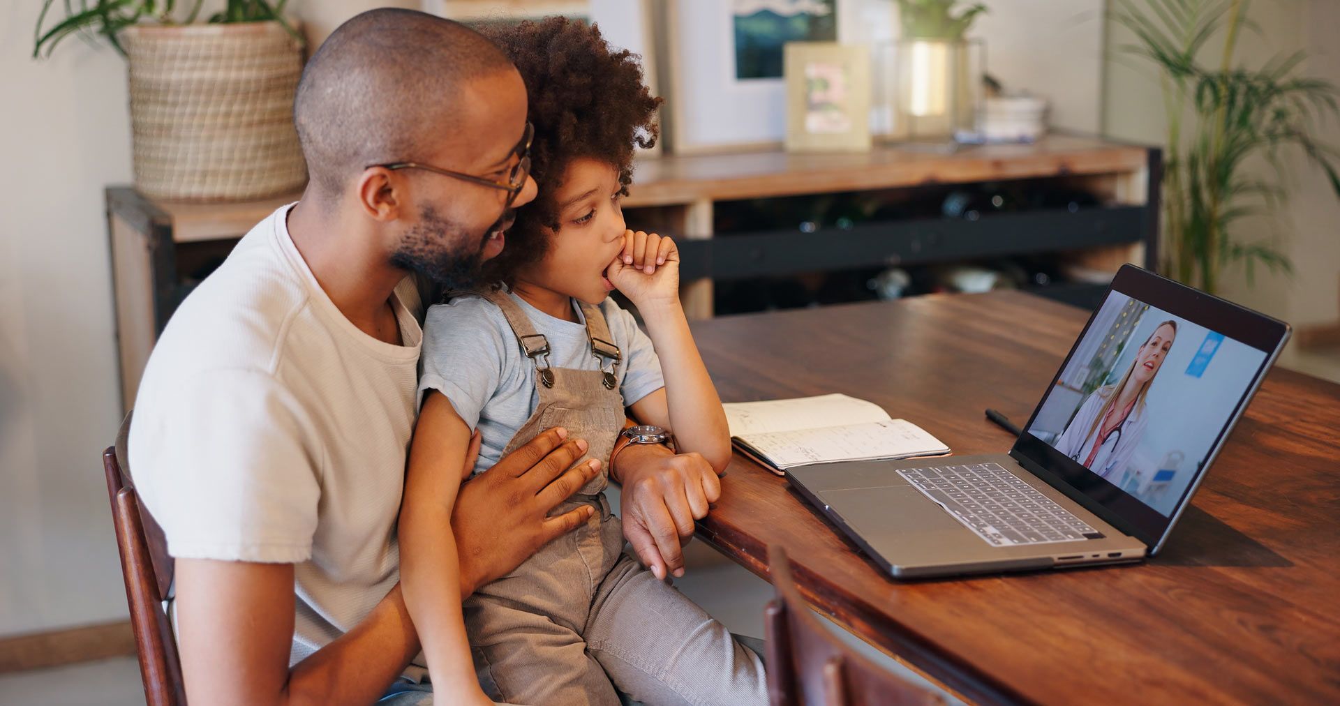 Father and child looking at laptop screen. They sit at a table, both are smiling.