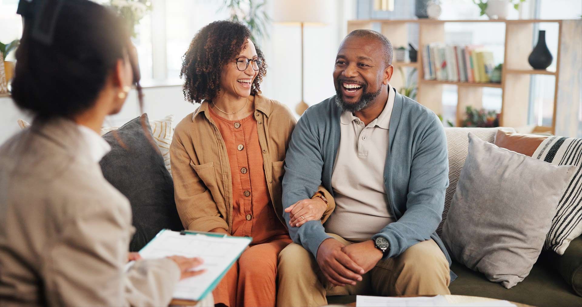 Couple on couch smiling at person holding clipboard. Indoors, warm lighting.