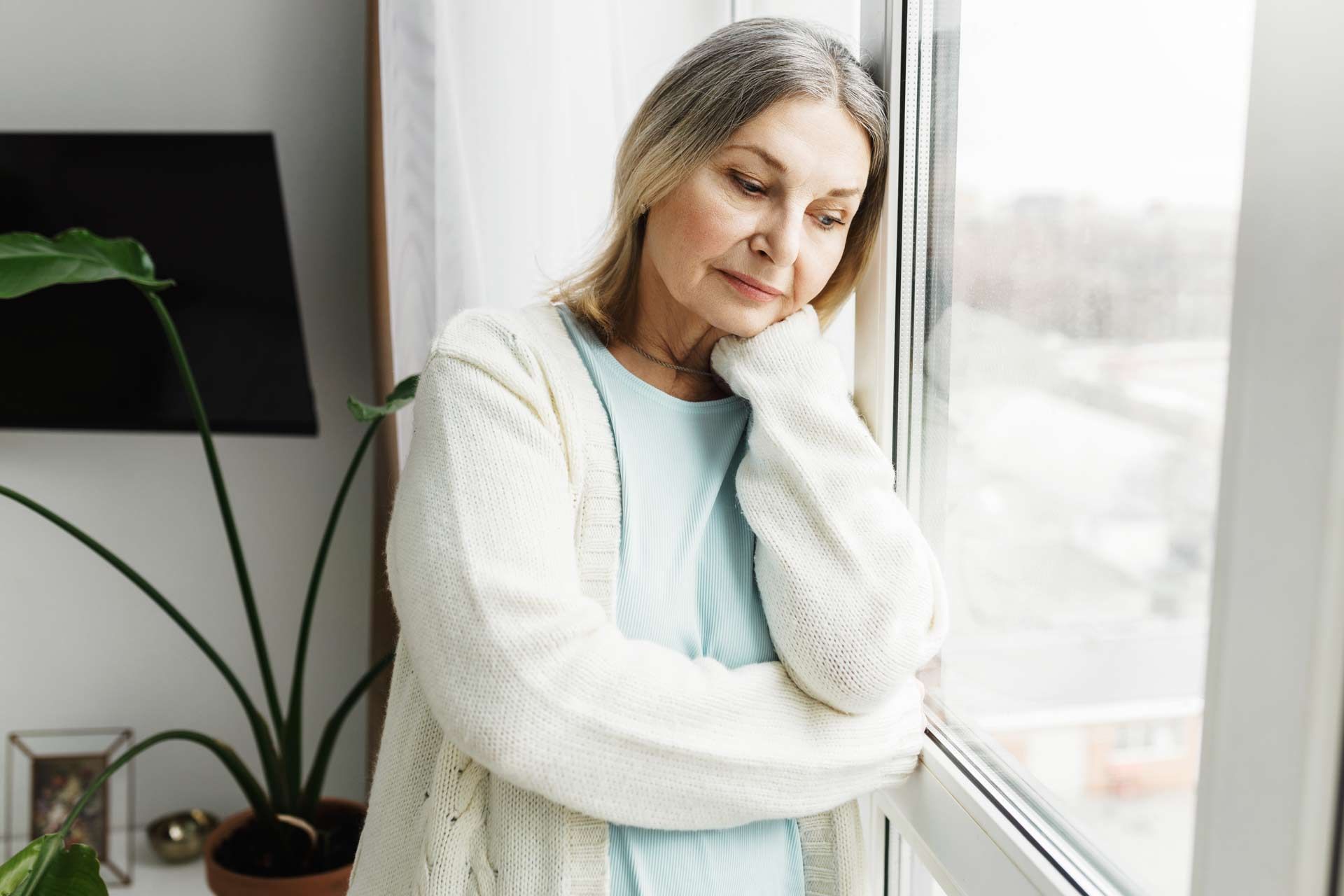 Woman with gray hair, looking out a window with a pensive expression, wearing a white cardigan.