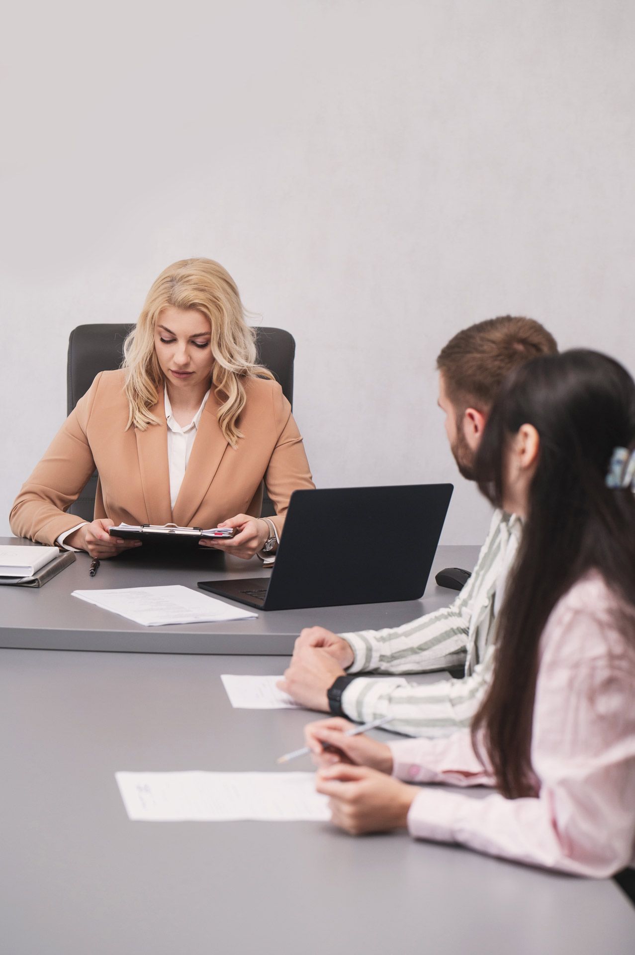 Woman at a table with a laptop, looking at a clipboard. Two people sit across from her.