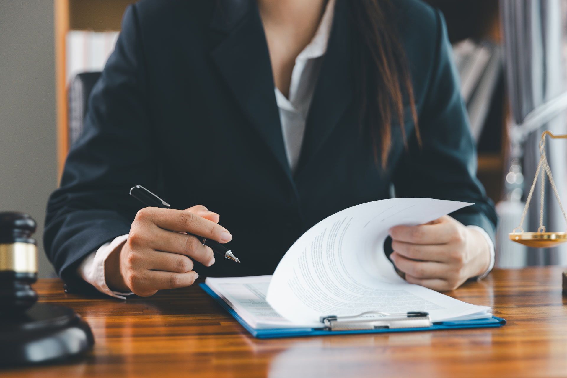 Woman in suit holding pen, reviewing documents at desk with gavel and scales of justice.