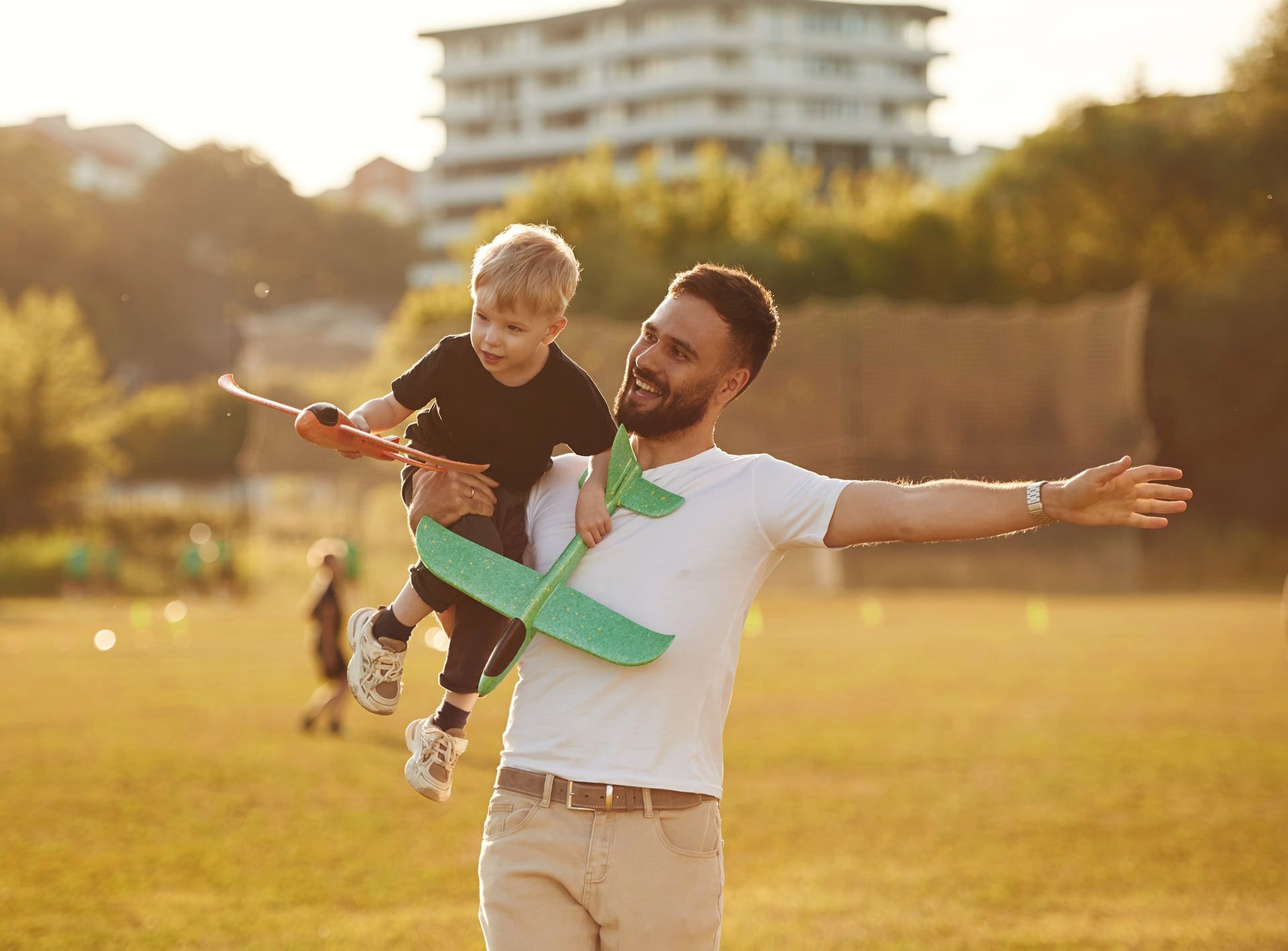 Man holding a child on his shoulders, both holding toy airplanes in a sunny park.