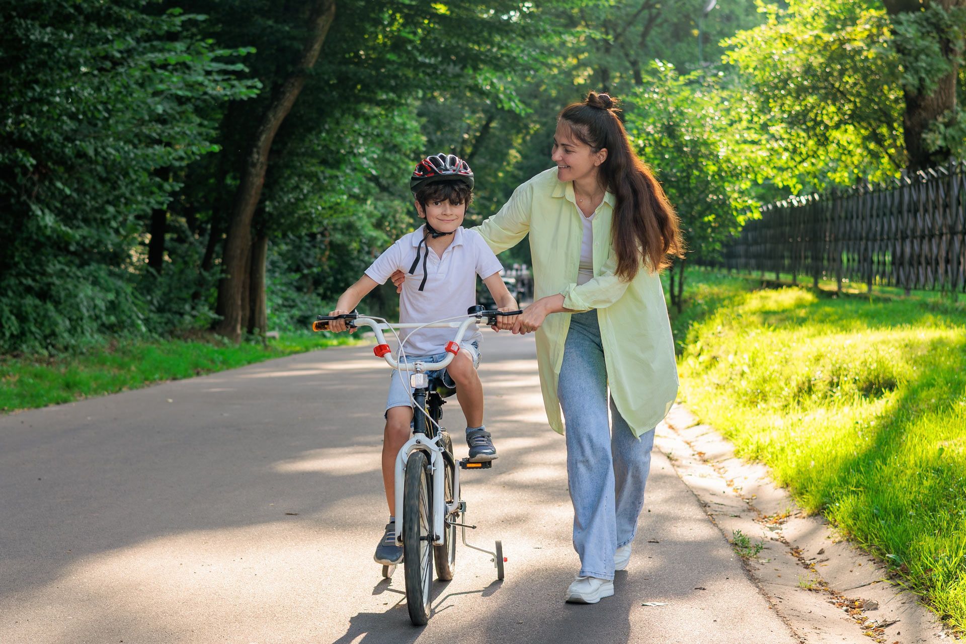 Woman helping child learn to ride a bike on a paved path in a park; sunny day.