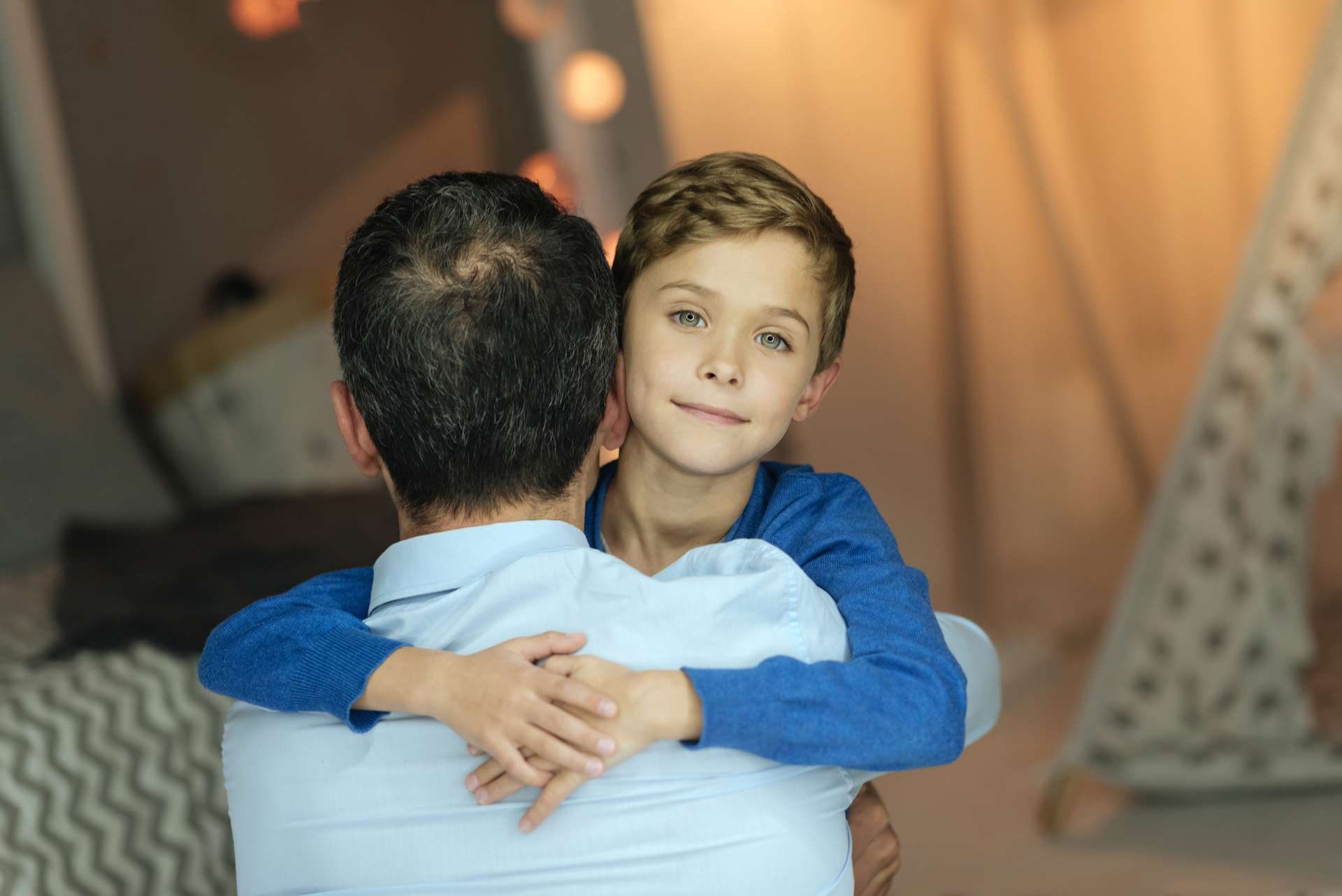 Boy hugging a man, both indoors. The boy smiles, wearing blue, the man's back is to the camera.