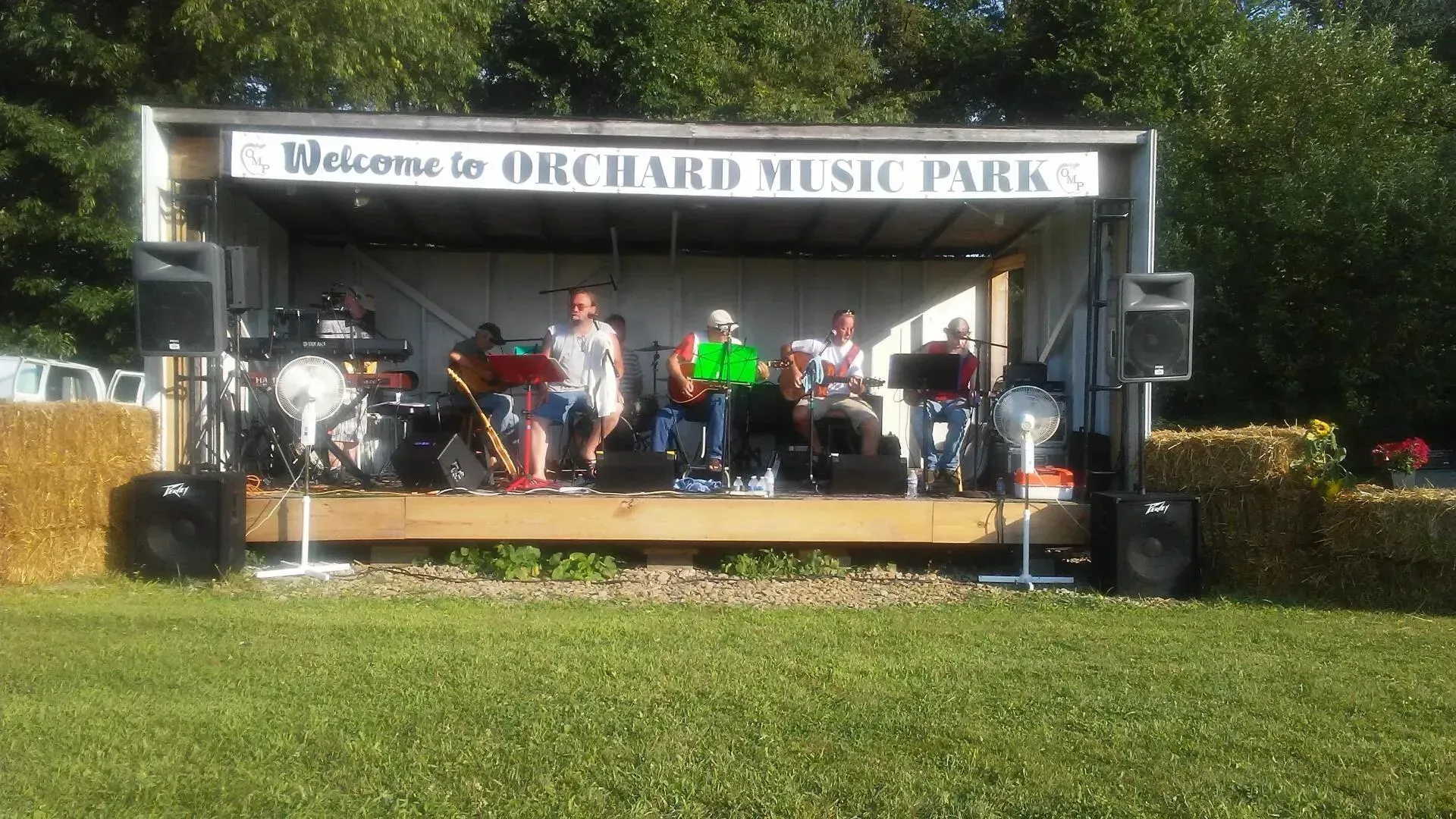 A group of people are playing instruments on a stage in an orchard music park.