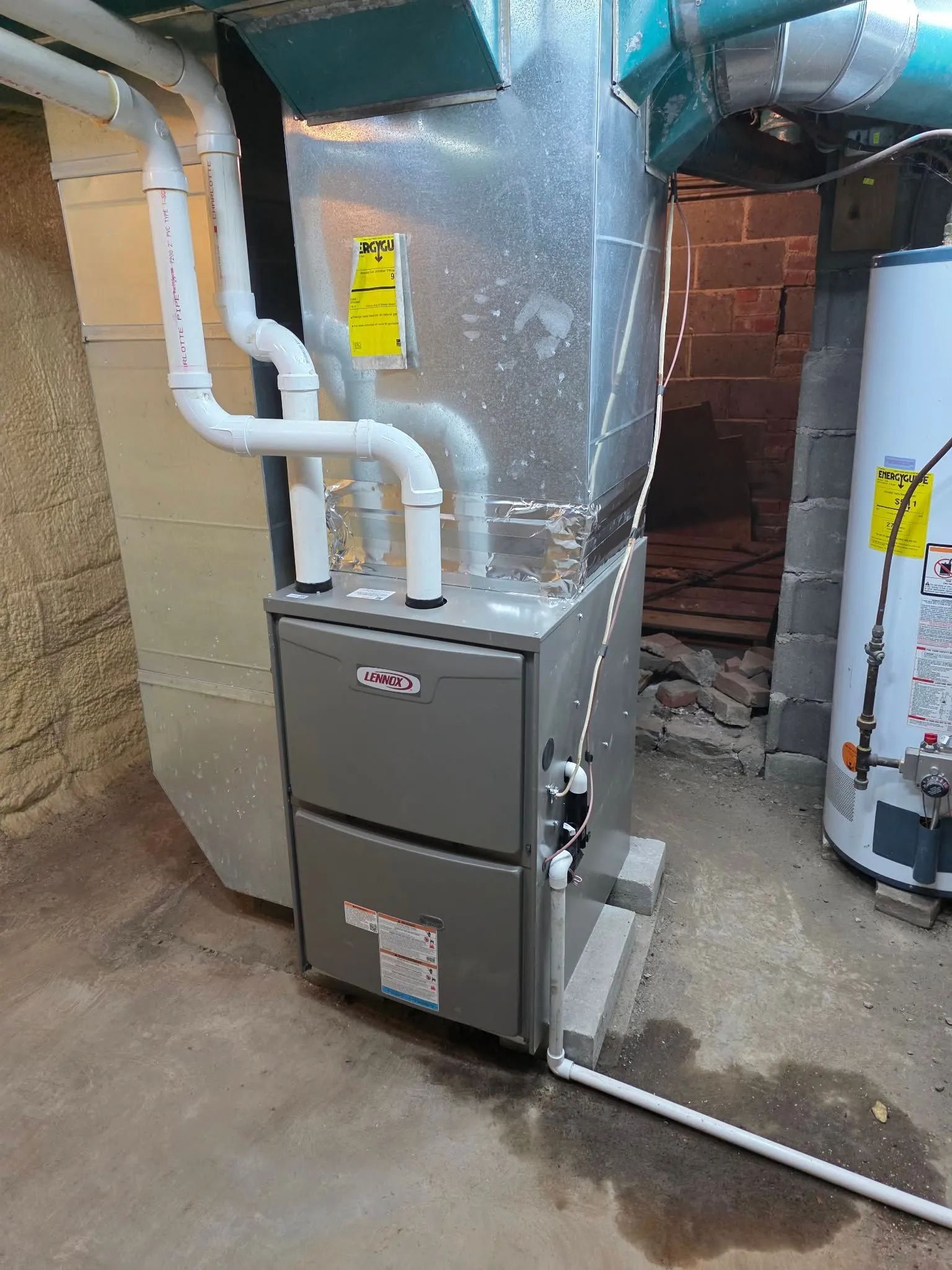 A grey furnace unit stands on a basement floor next to a white water heater, with white PVC piping running along the side.