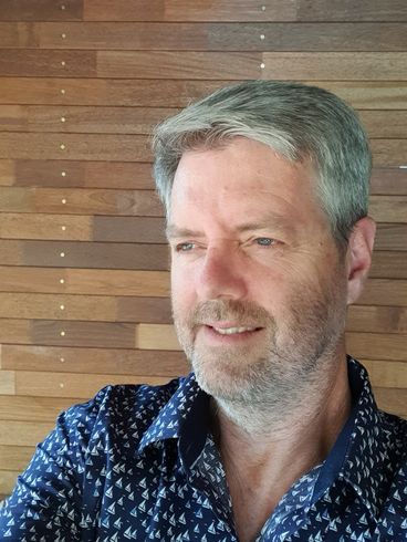 A man with a beard and a blue shirt is smiling in front of a wooden wall.