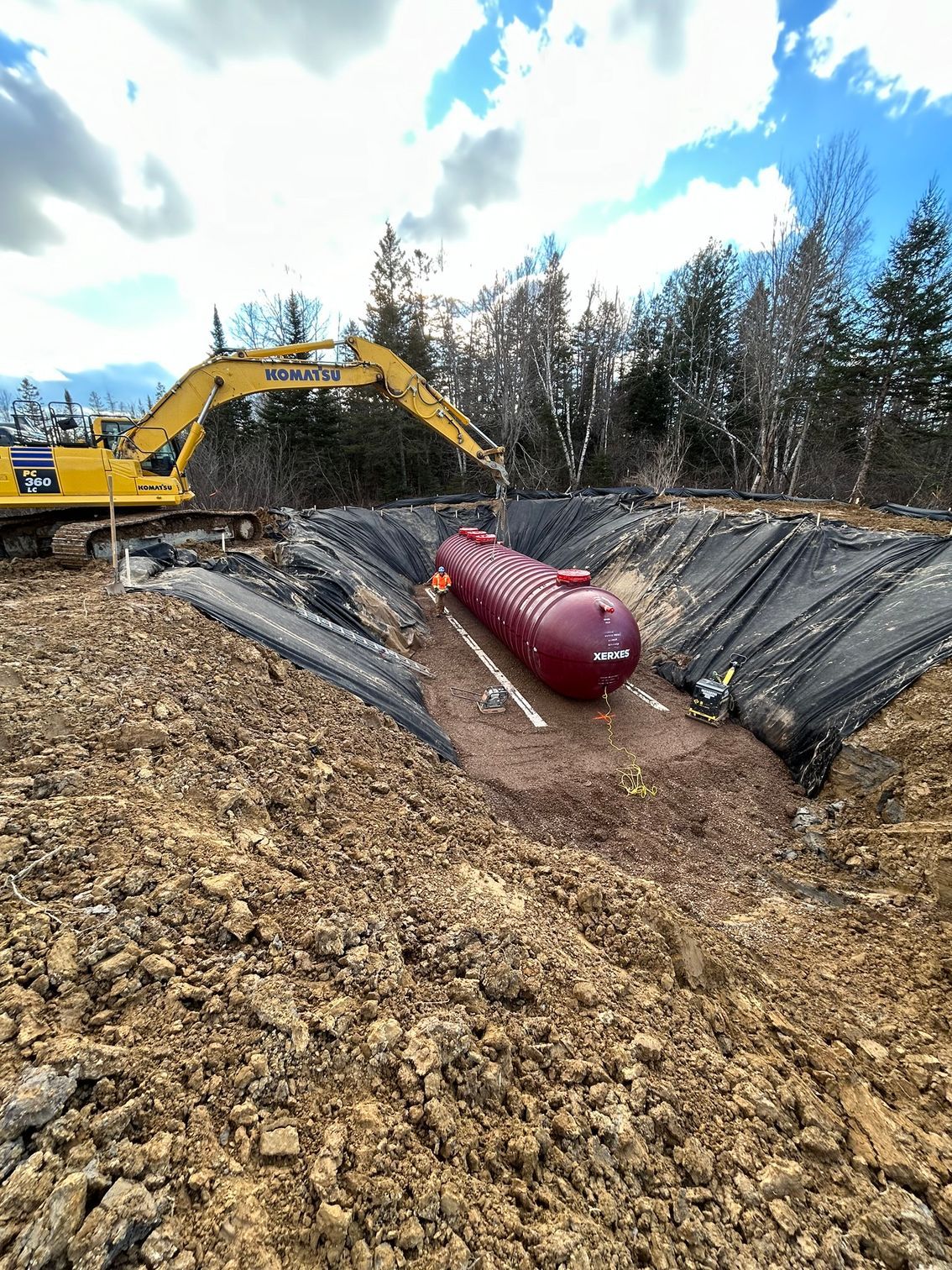 A red tank is sitting in the middle of a dirt field next to a yellow excavator.