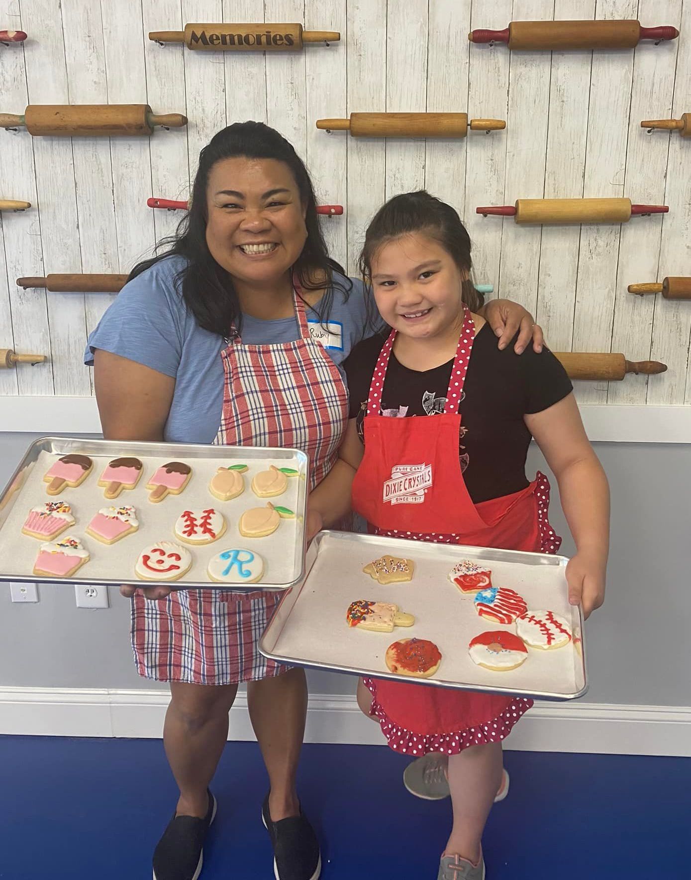 Woman and girl in aprons holding trays of decorated cookies, smiling. Rolling pins background.