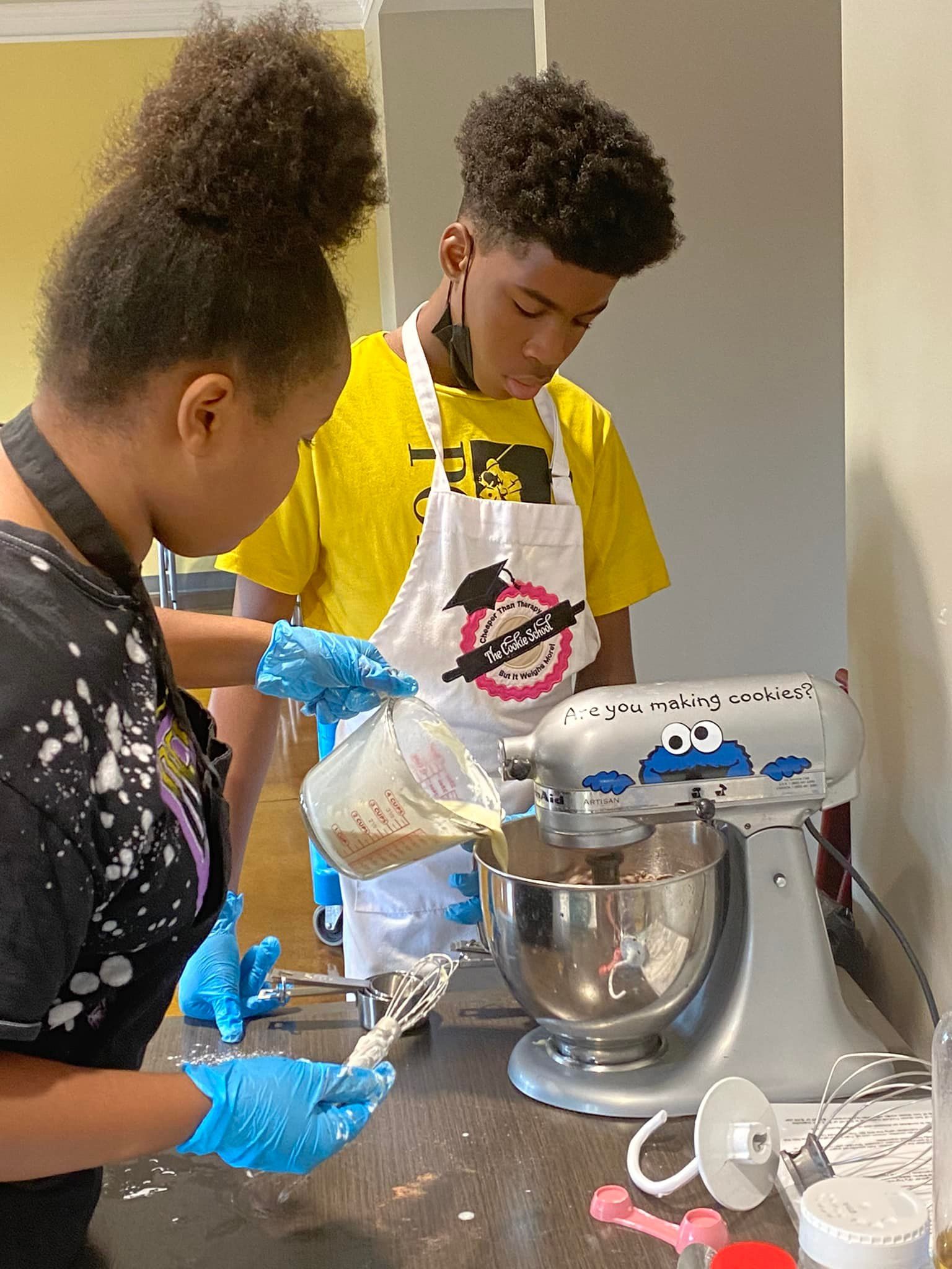 Two people baking together, pouring ingredients into a mixer. They wear aprons and gloves in a kitchen.
