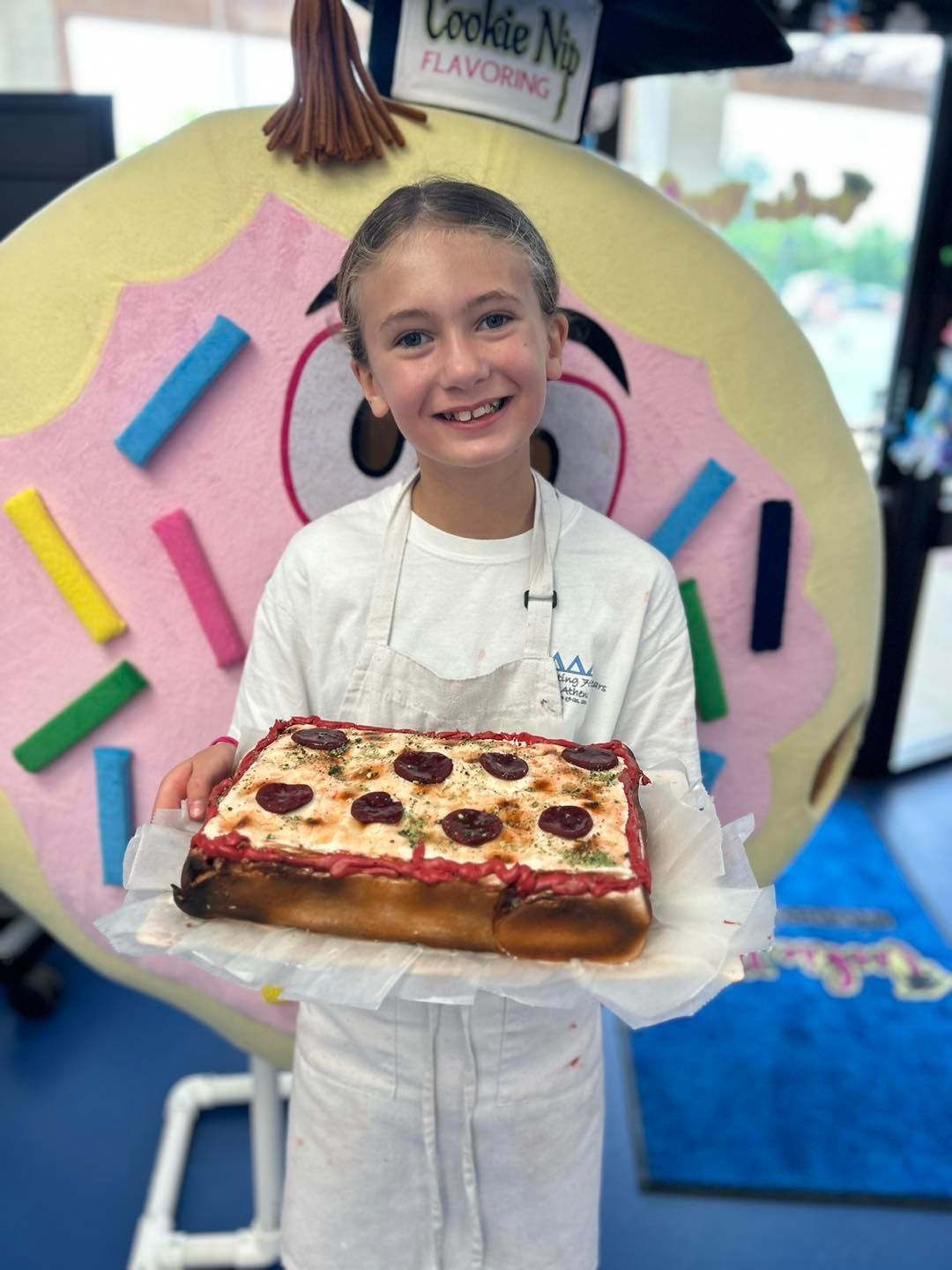 Girl holding a rectangular pizza, smiling, standing in front of a donut-shaped mascot.