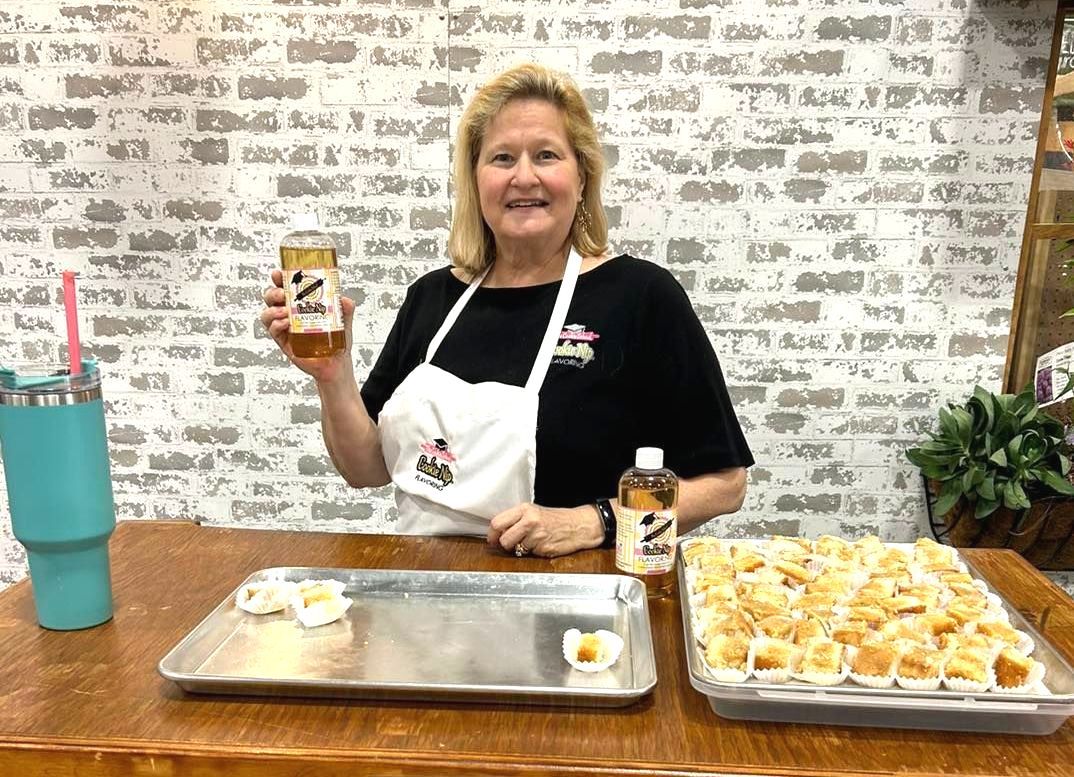 Woman in apron holds up a jar and can of product in front of a tray of baked goods and a brick wall.