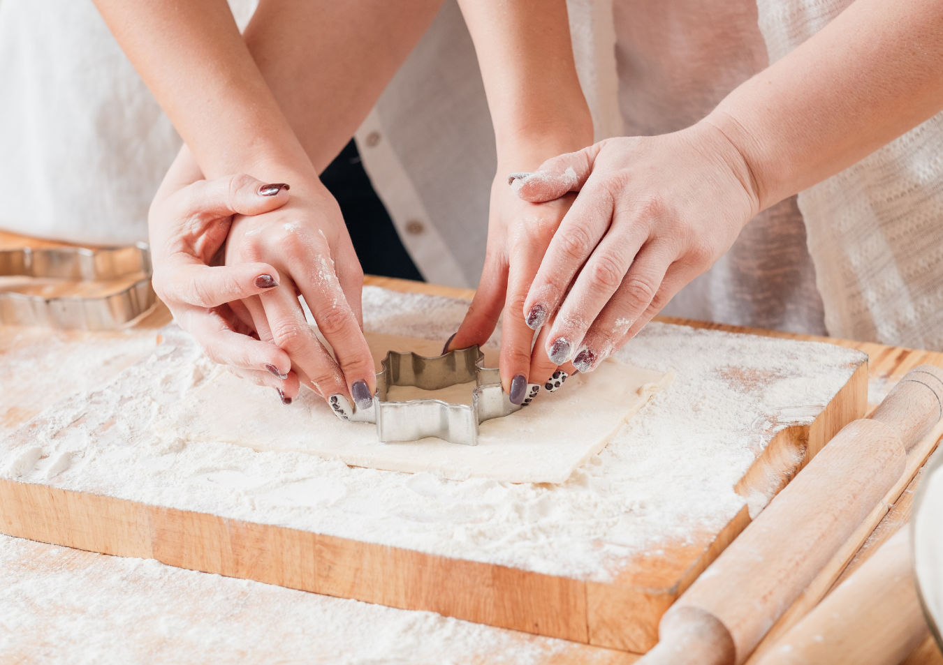 Hands using a cookie cutter on a flour-covered wooden board, rolling pins visible.