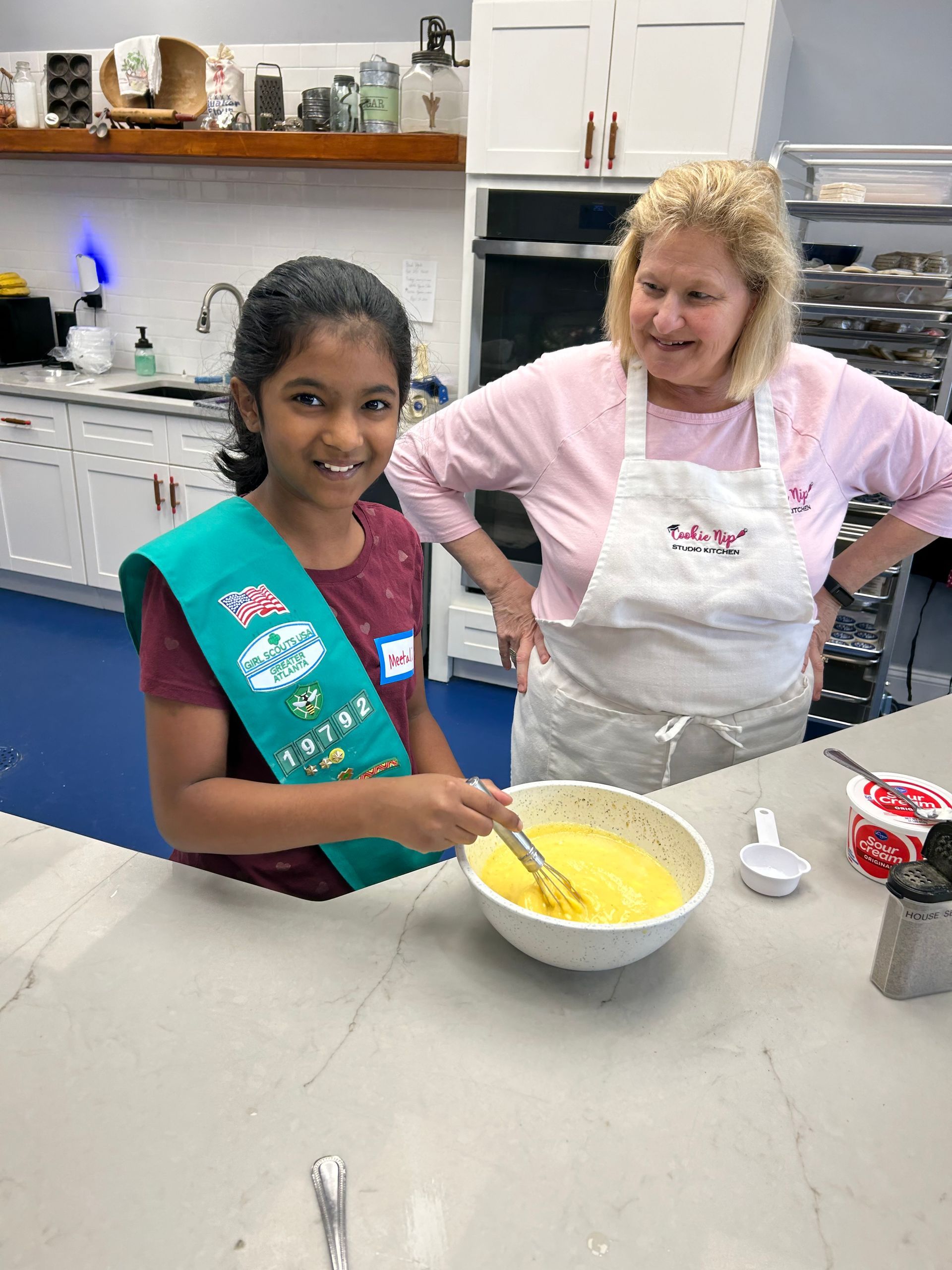 Girl Scout stirring batter in a kitchen with an instructor; smiling and wearing a sash and apron.