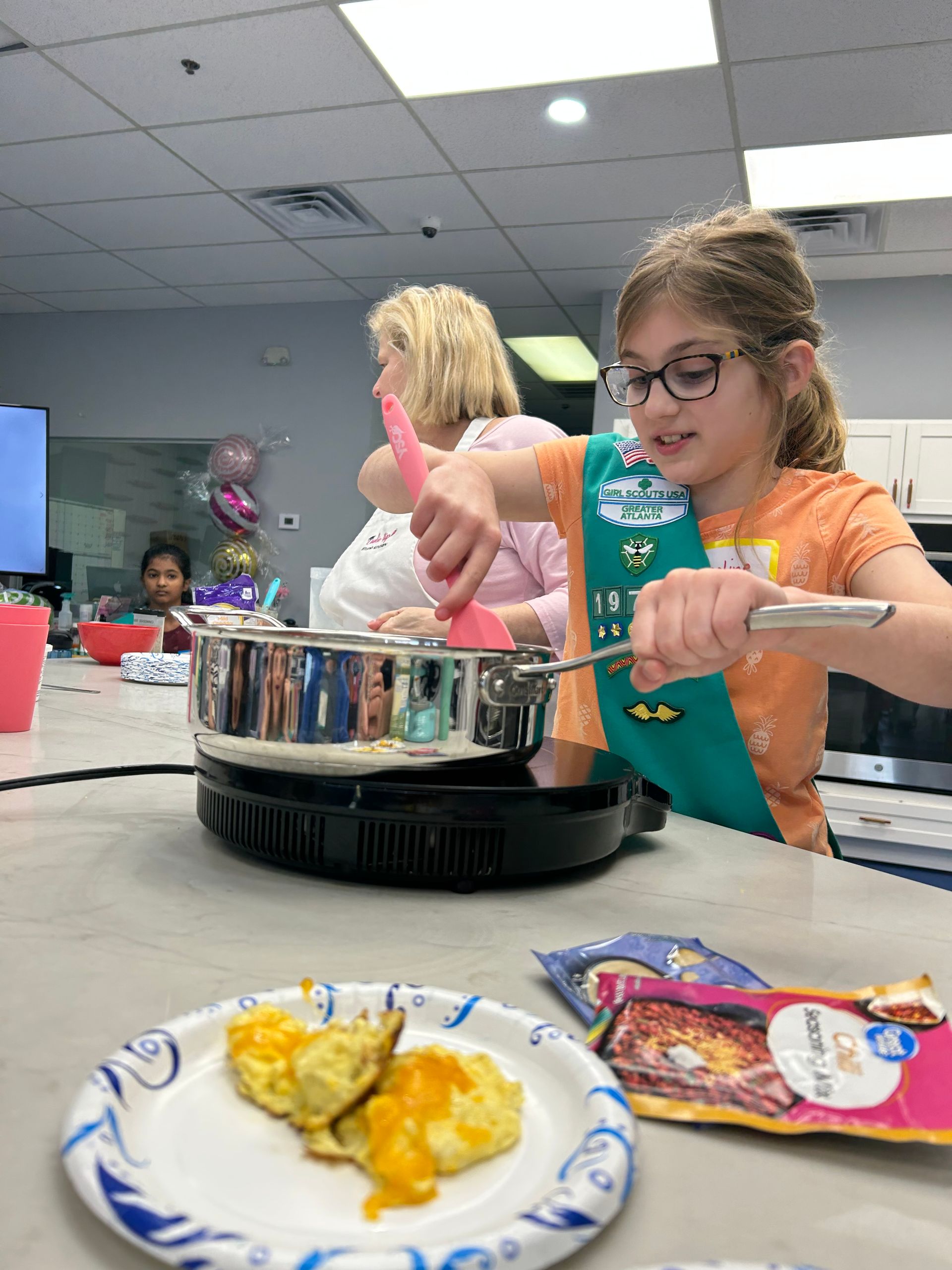 Girl Scout cooking at a stove, stirring food. A plate of food in the foreground and woman in the background.