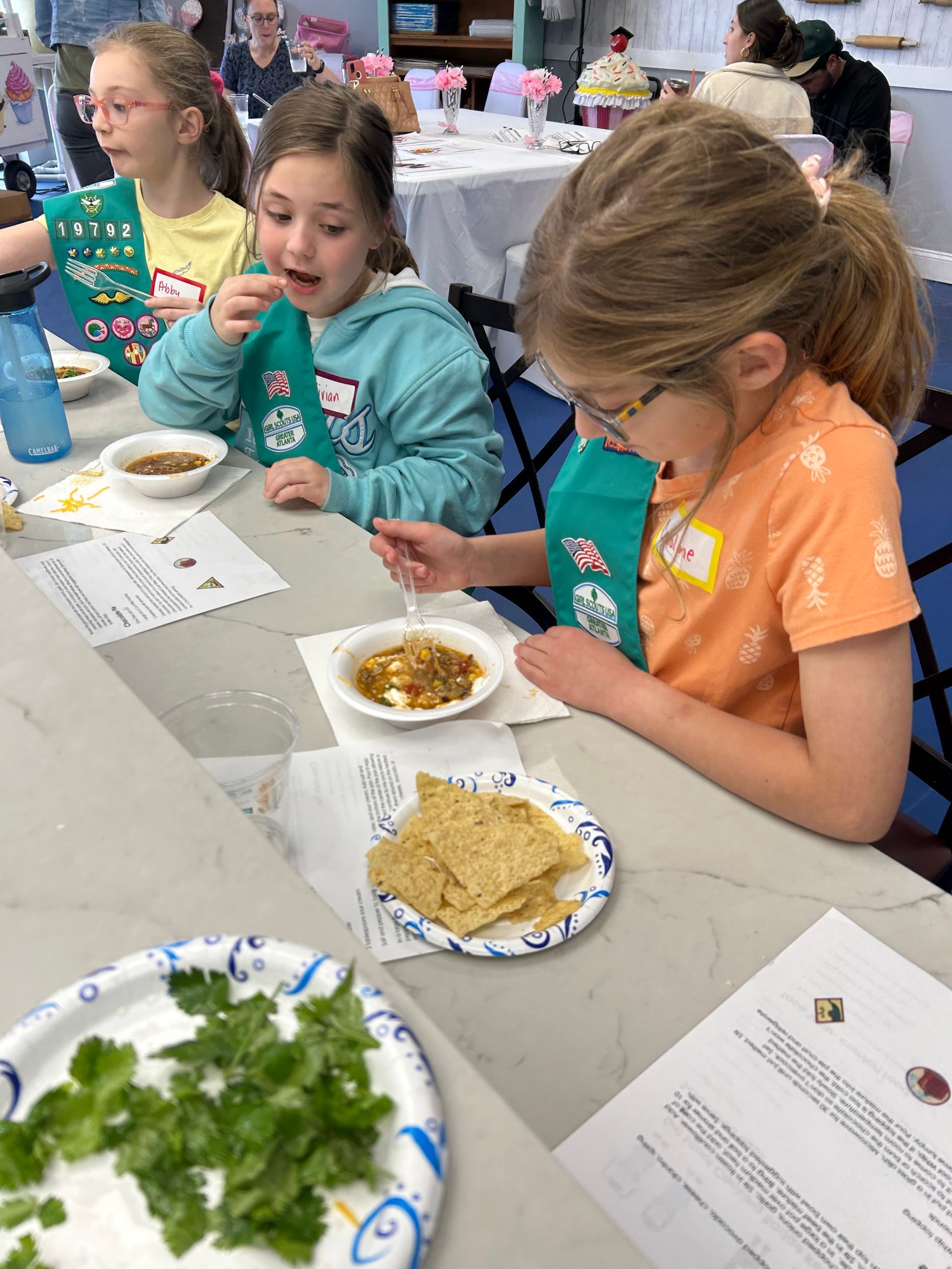 Three girls tasting food at a table. Bowls of food, crackers, and parsley are visible.