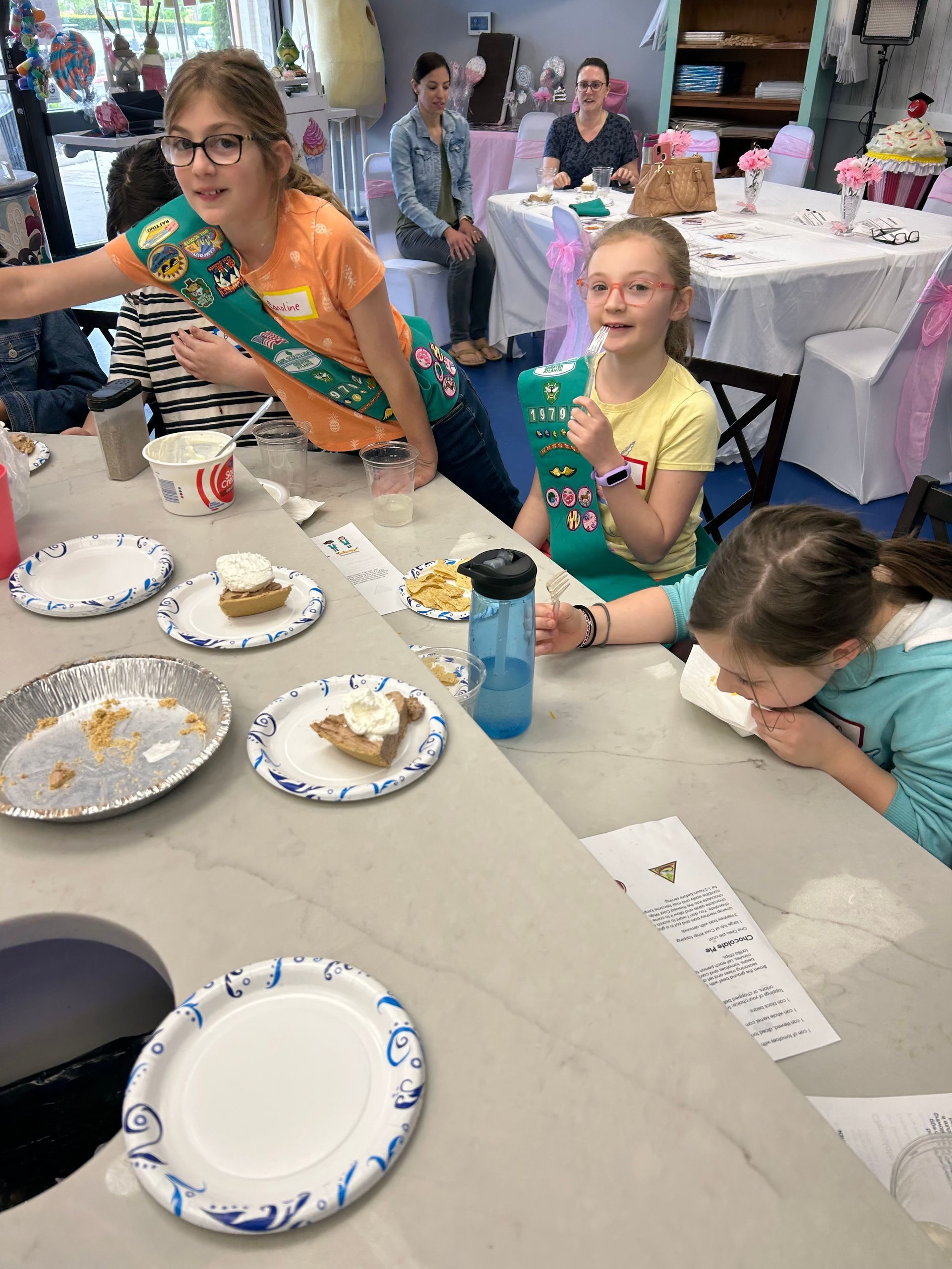 Girl Scouts at a table eating treats. Two wear green sashes, one is leaning over, another eating, and a third eating.