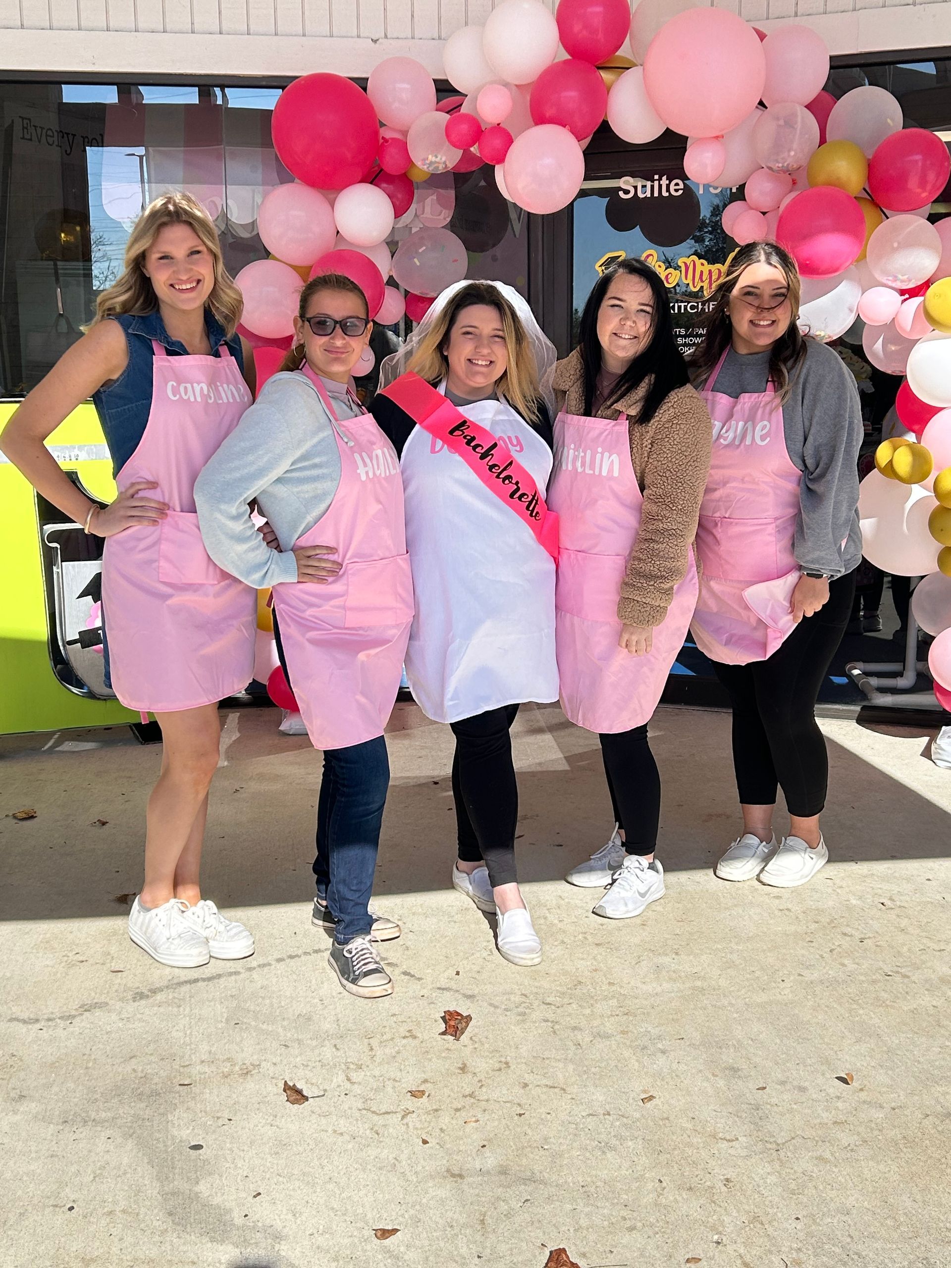 Five women in pink aprons and a bride-to-be with a sash pose outside a store, surrounded by pink balloons.