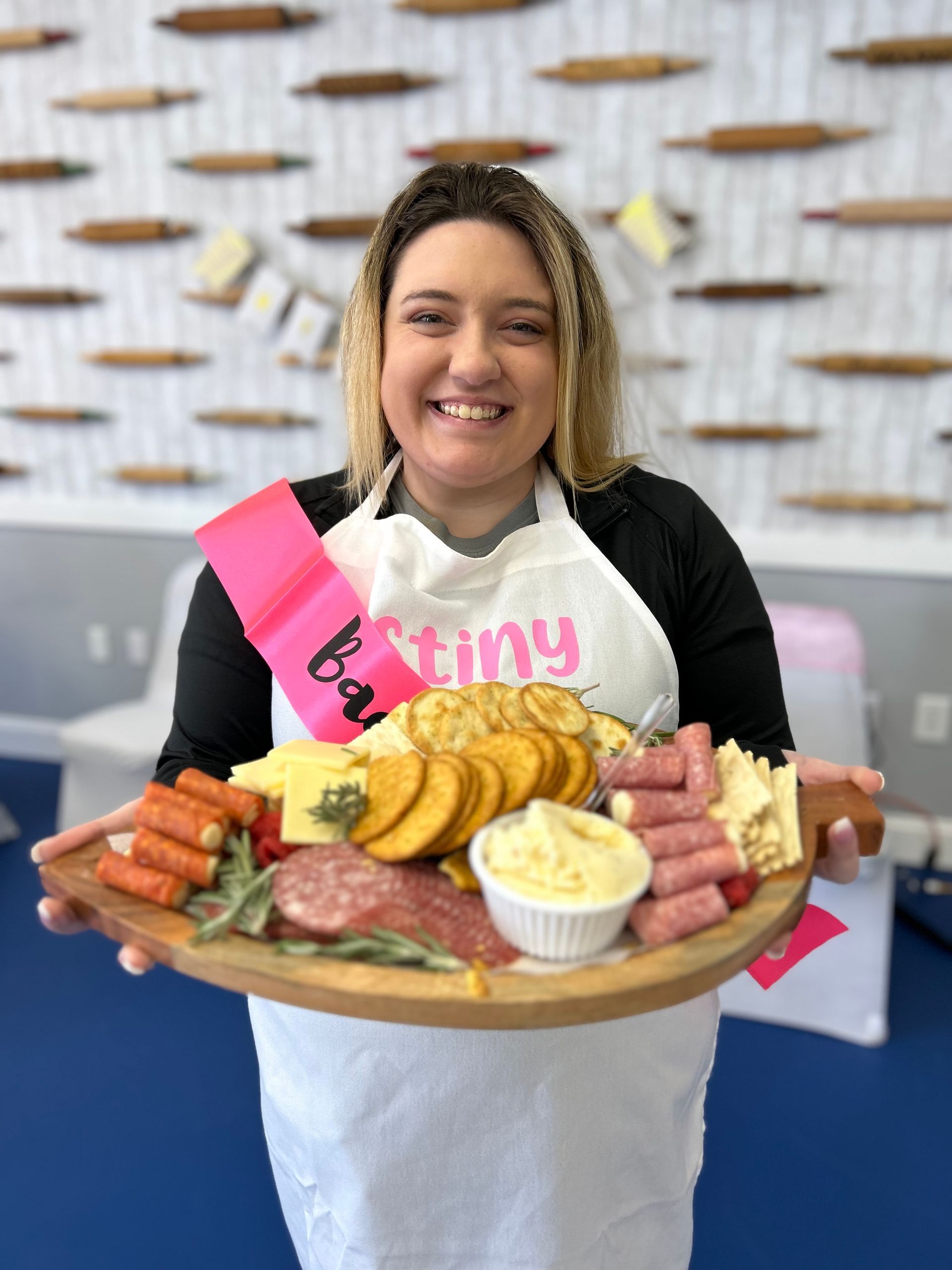 Woman smiles, holding a charcuterie board with snacks, wearing a