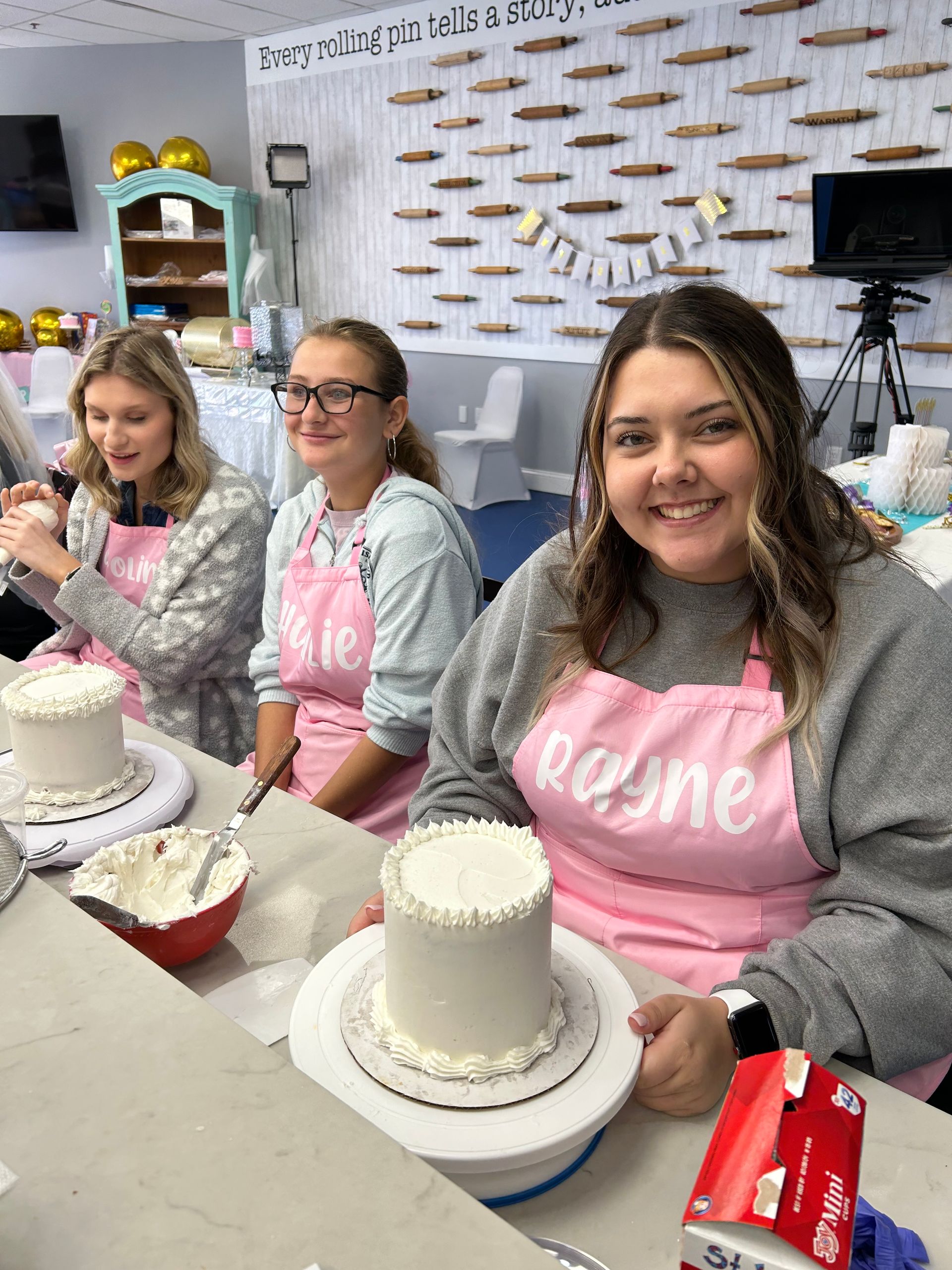 Three people in pink aprons decorating cakes at a workshop, smiling and focused.