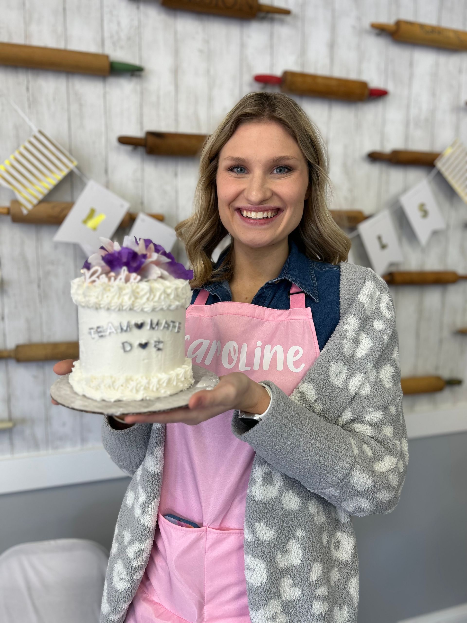 Woman holding a white cake with purple flowers. She wears a pink apron and stands in front of rolling pins.