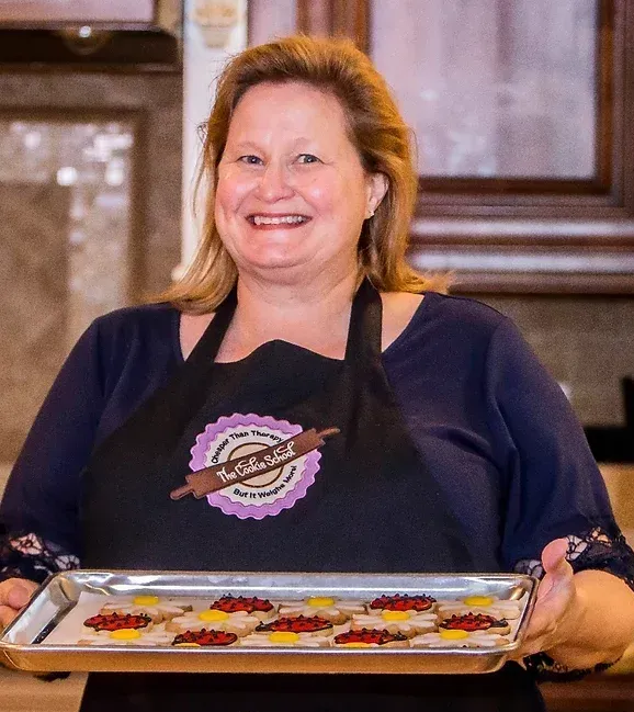 Woman holding a tray of decorated cookies, wearing an apron, smiling.