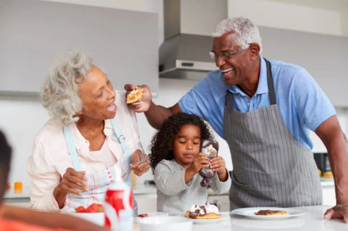 Family in kitchen making pancakes, smiling. Grandparents and a child are seen.