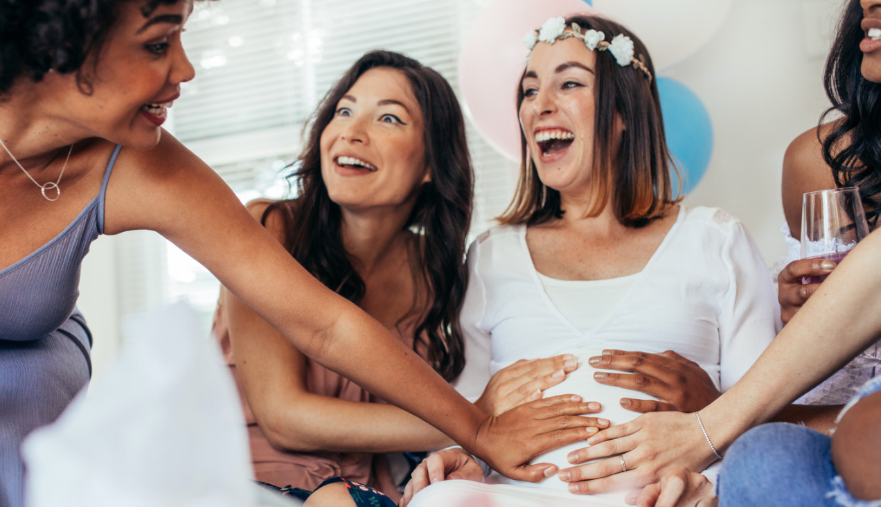 Women laughing and touching a pregnant woman's belly at a baby shower.