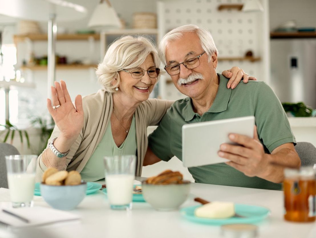 Older couple video chatting on a tablet, waving hello. They're at a table with food and drinks indoors.