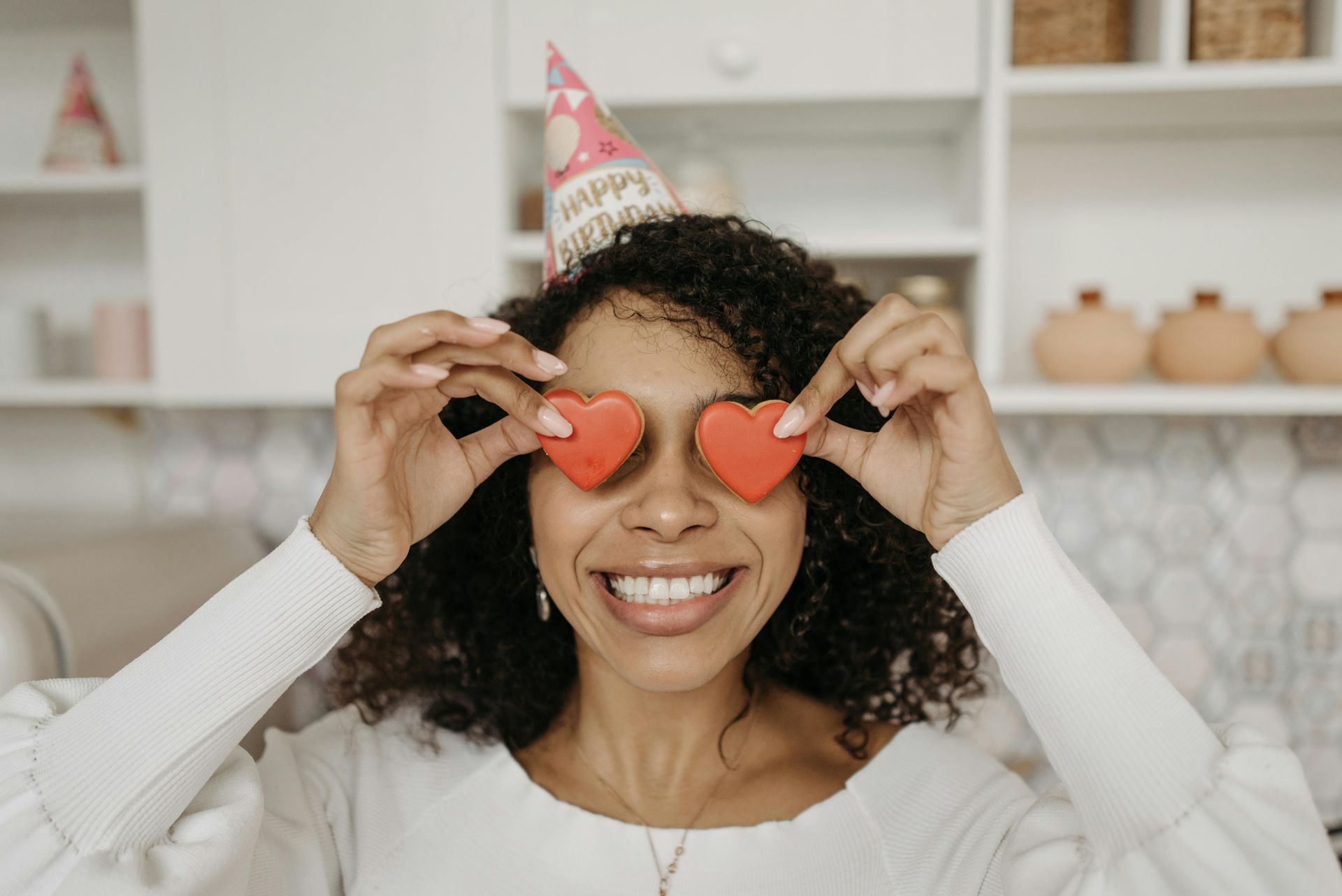 Woman holding red heart-shaped cookies over her eyes, wearing a party hat, smiling brightly in a kitchen.