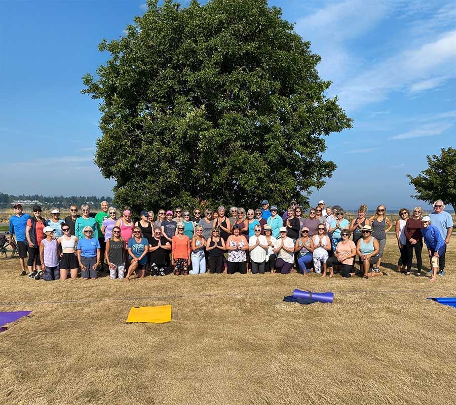 A large group poses for a photo on a dry, grassy field in front of a tall tree, most with hands in a prayer pose.