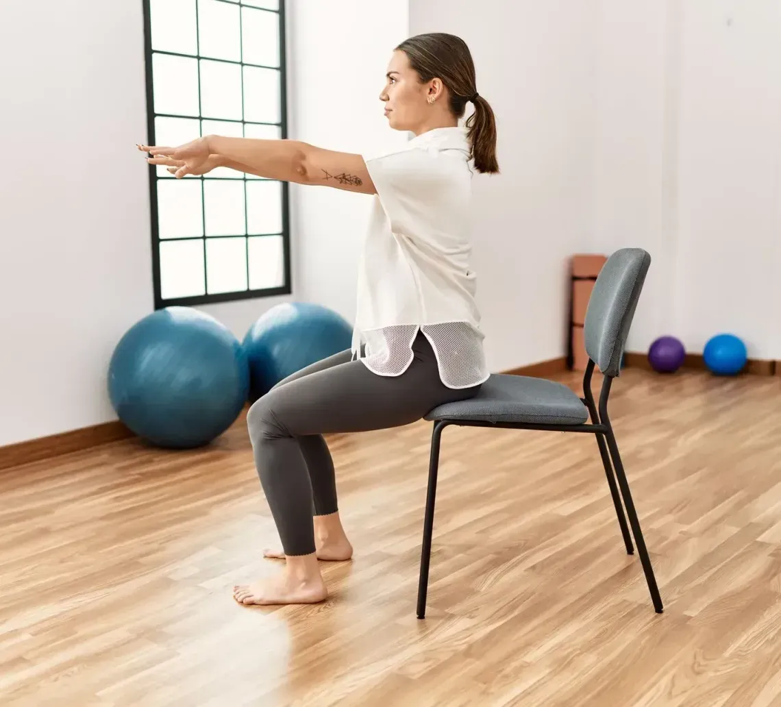 A person kneeling on a yoga mat performs a chair-supported child's pose in a light-filled room.