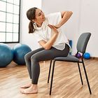 A person sits on a chair in a studio, performing a torso-twisting stretch with palms pressed together.